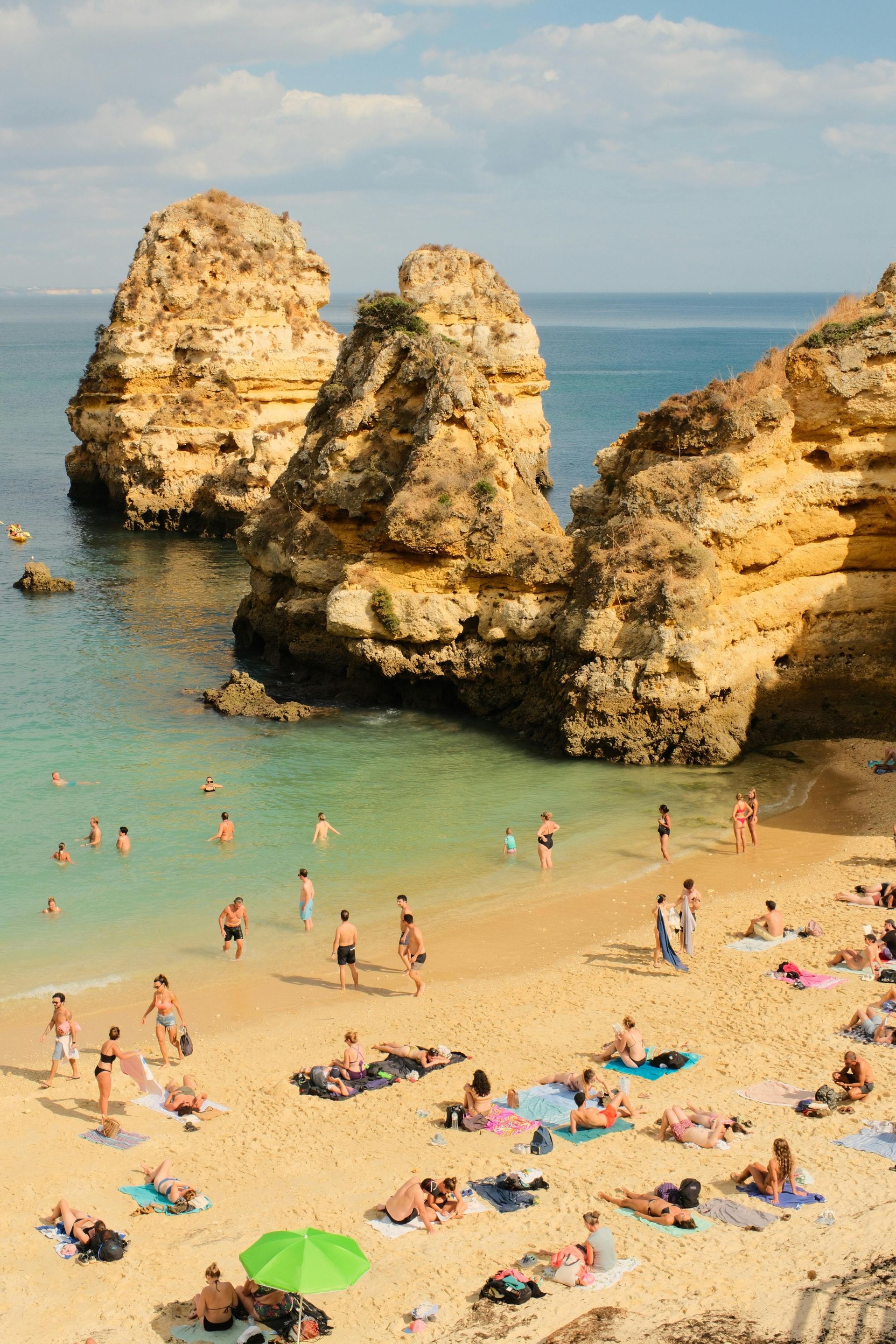 People relax and swim at a sandy beach enclosed by large, golden limestone rock formations under a bright blue sky.