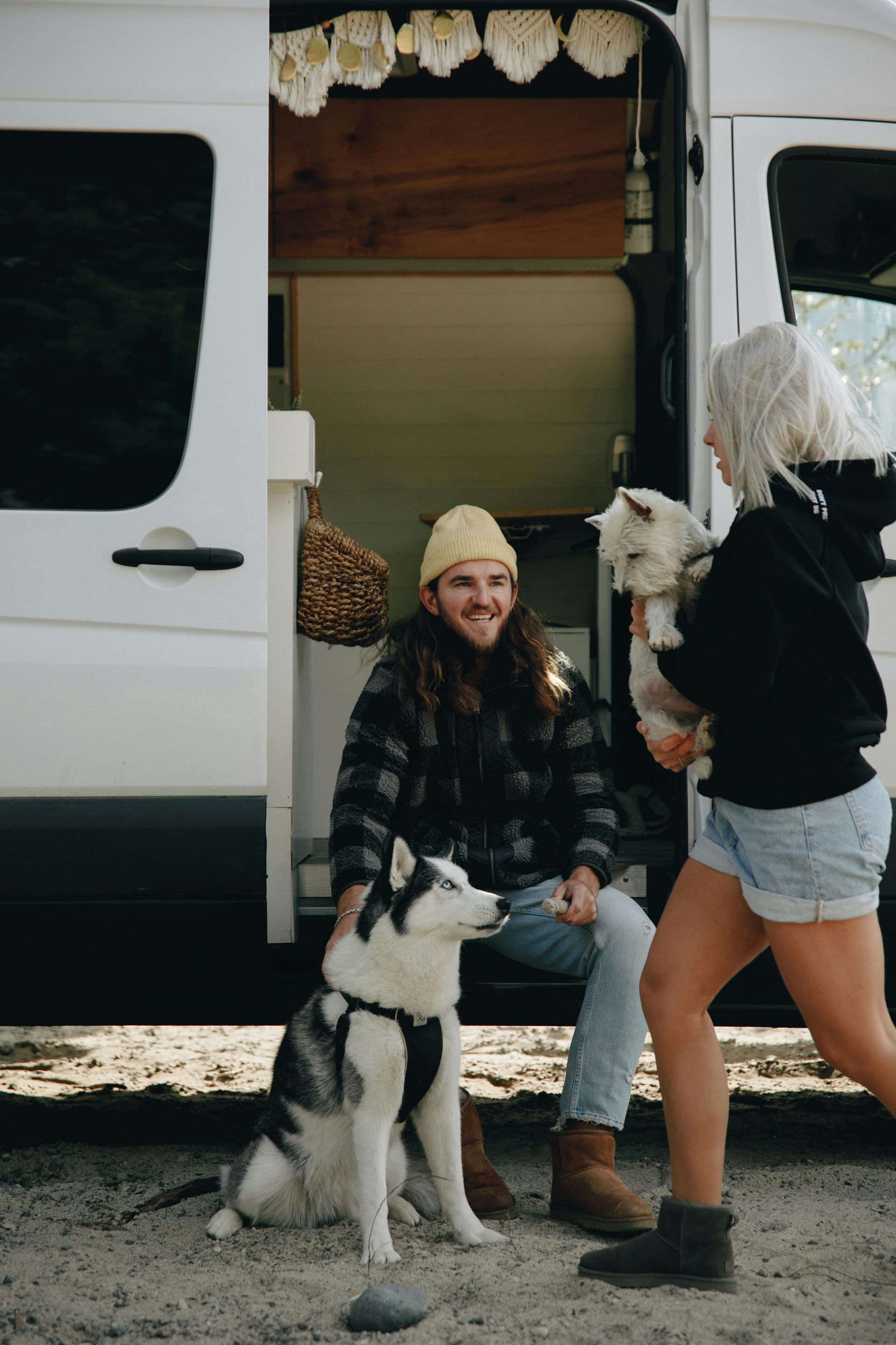 A smiling person sits in a van doorway petting a husky, while another person stands nearby holding a small dog.
