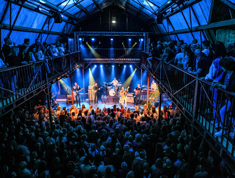 Band performs on stage in a dark barn-like venue, illuminated by blue and yellow lights; audience watches.