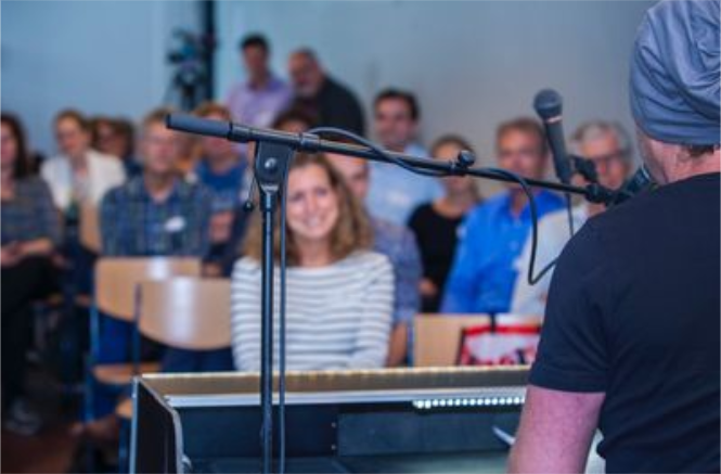 Person plays keyboard and sings to an audience seated in a room, with smiling faces in the audience.