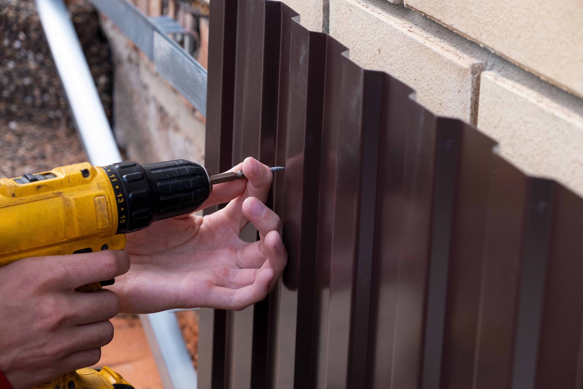Person using a yellow drill to screw a panel into a brick wall; brown metal panel and brown screw are visible. Person using a yellow drill to screw a panel into a brick wall; brown metal panel and brown screw are visible.
