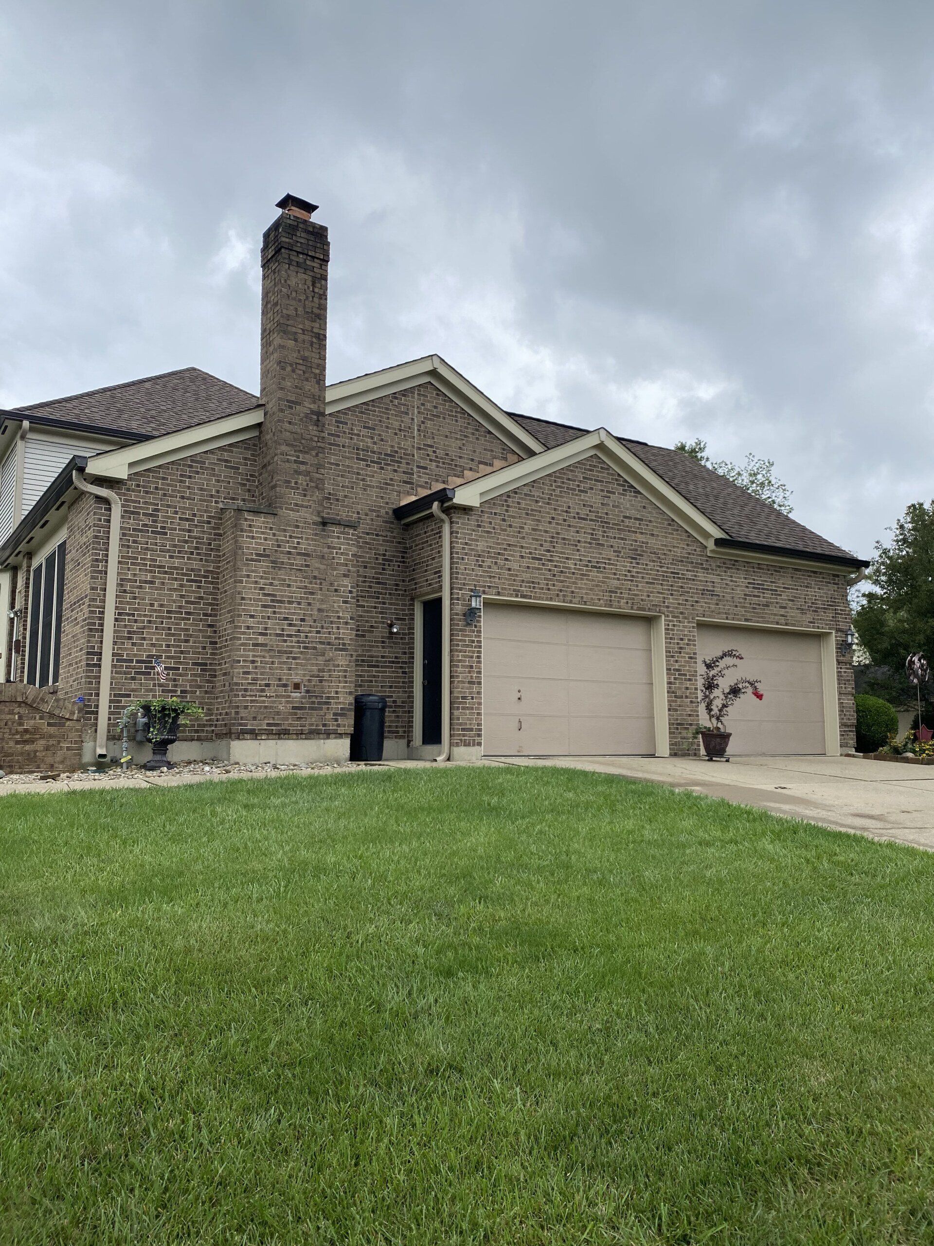 Roofing Replacement Services Near You Brick suburban house with a tall chimney, beige garage, and green lawn under cloudy skies