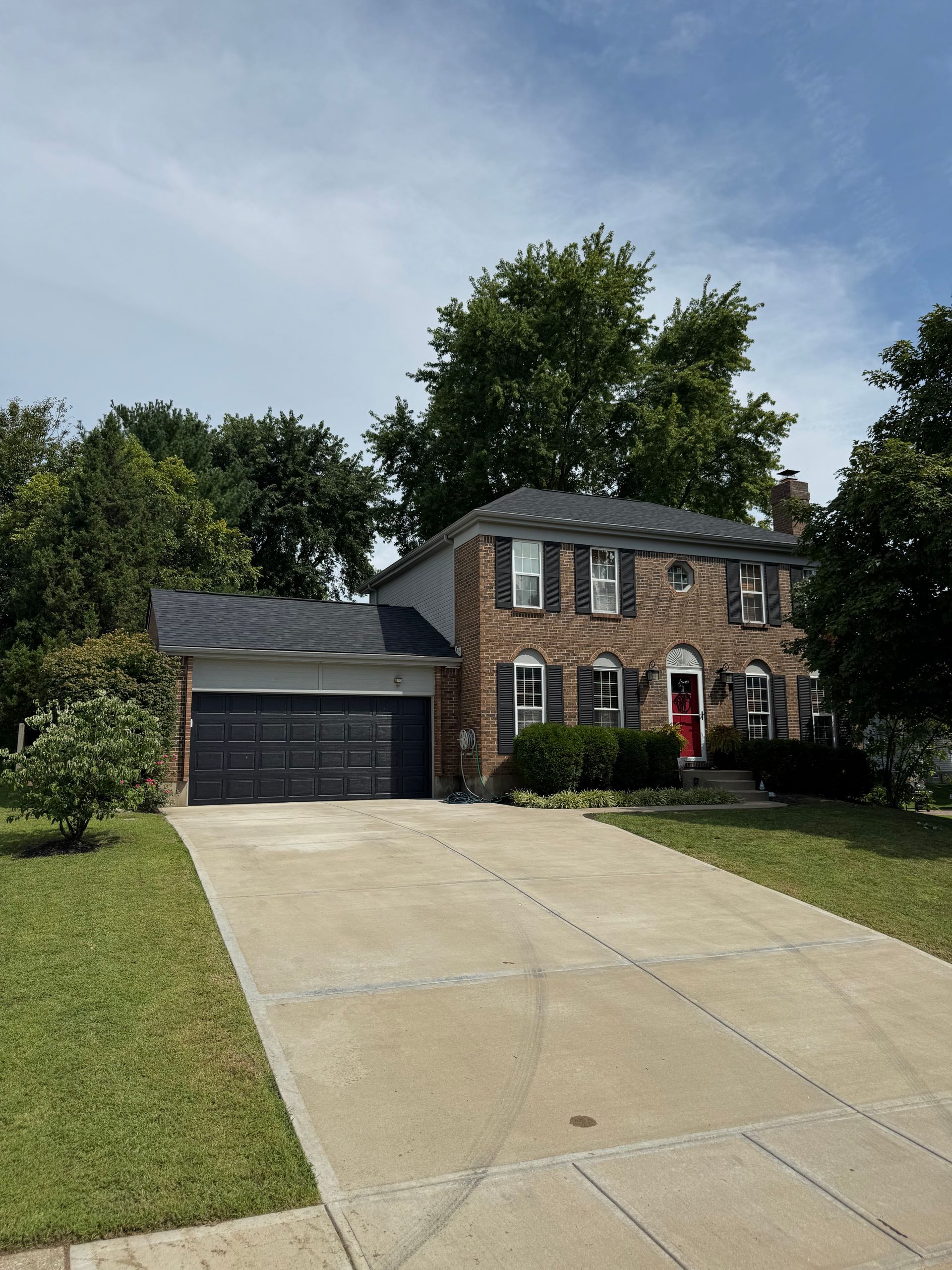 Roof Replacement Milford, OH Two-story brick house with black garage, red front door, and long driveway under a blue sky