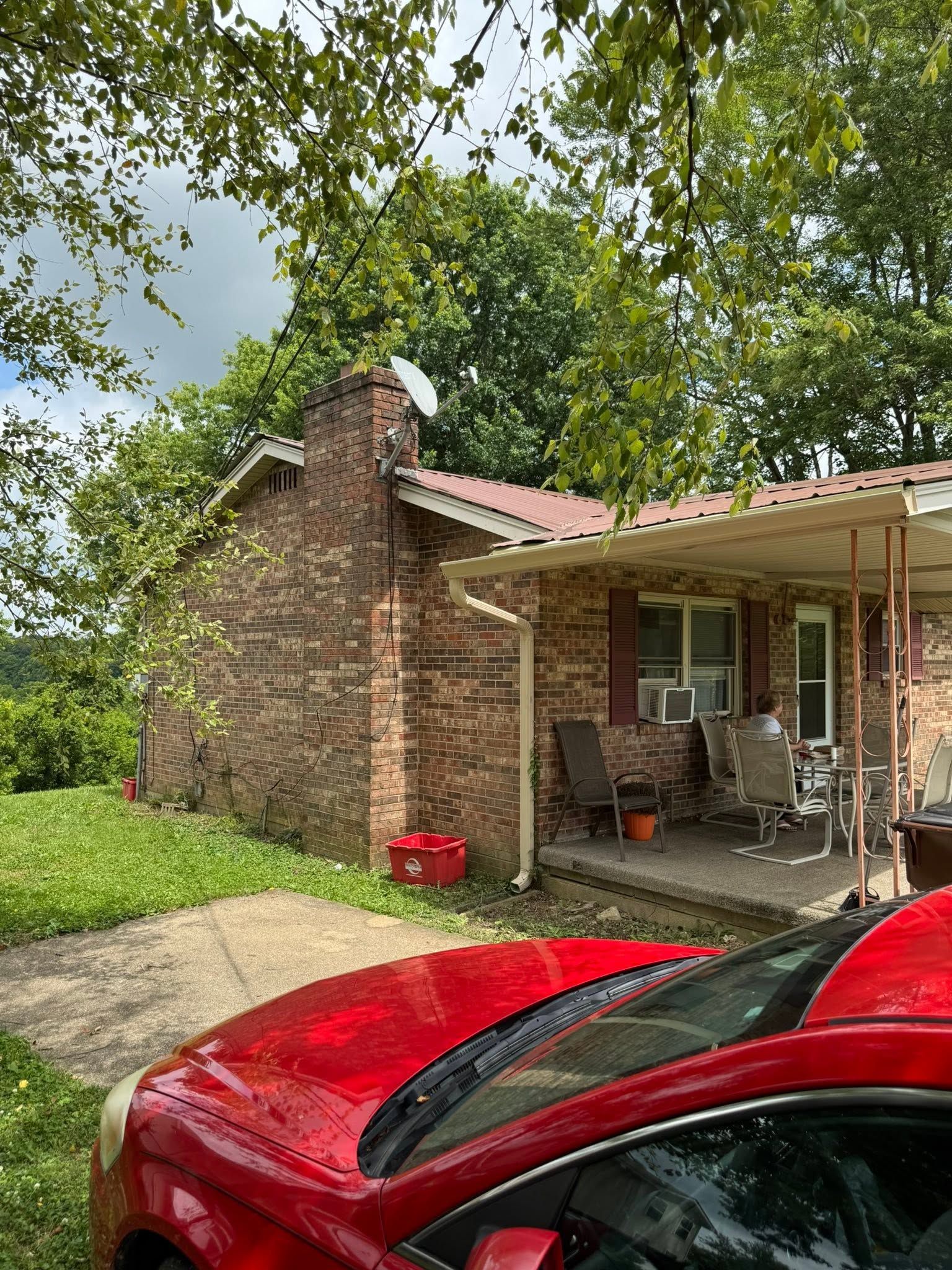 Red car parked by a brick house with a covered porch under leafy trees