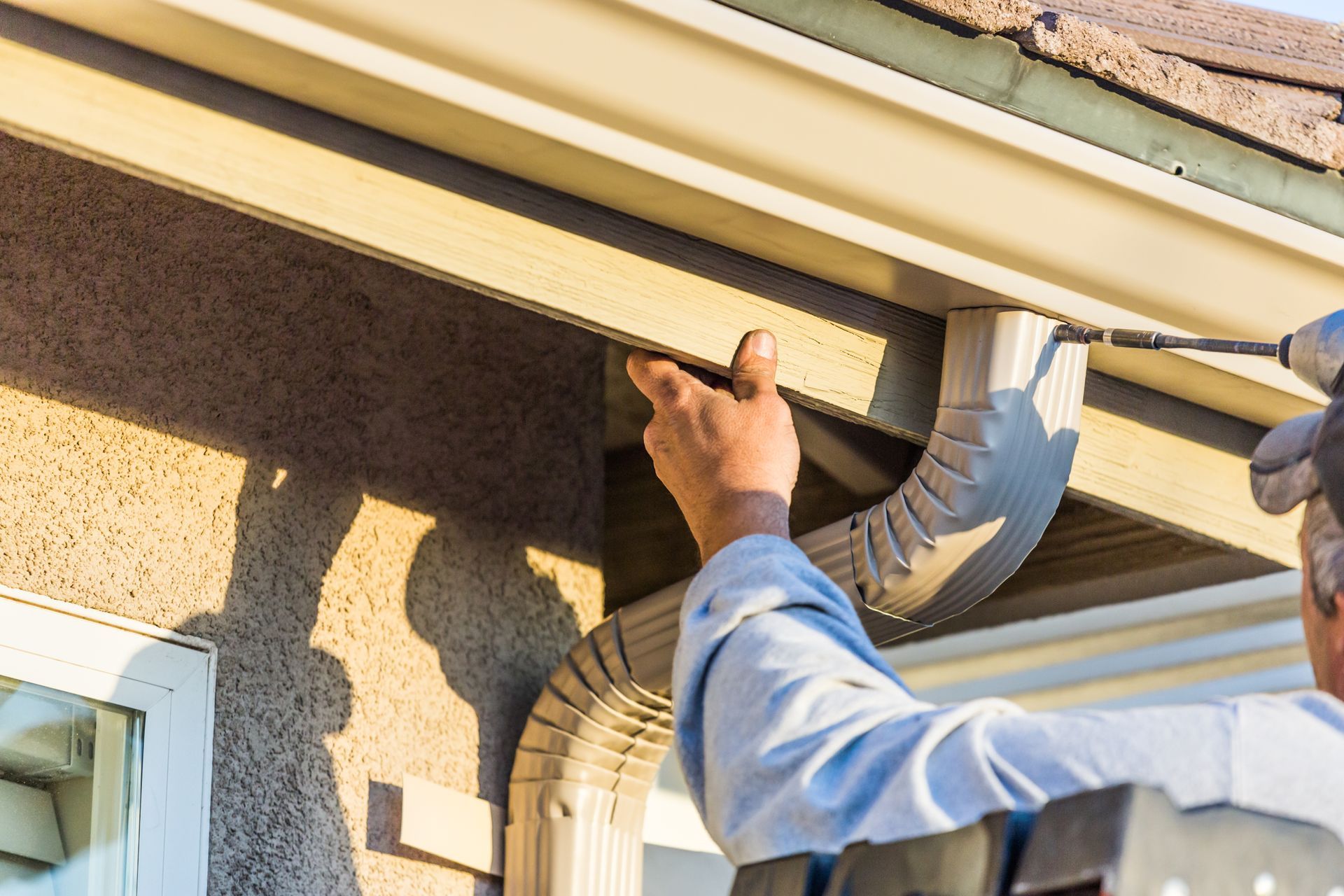 A contractor installing a seamless aluminum gutter and downspout on a home's roofline with a drill