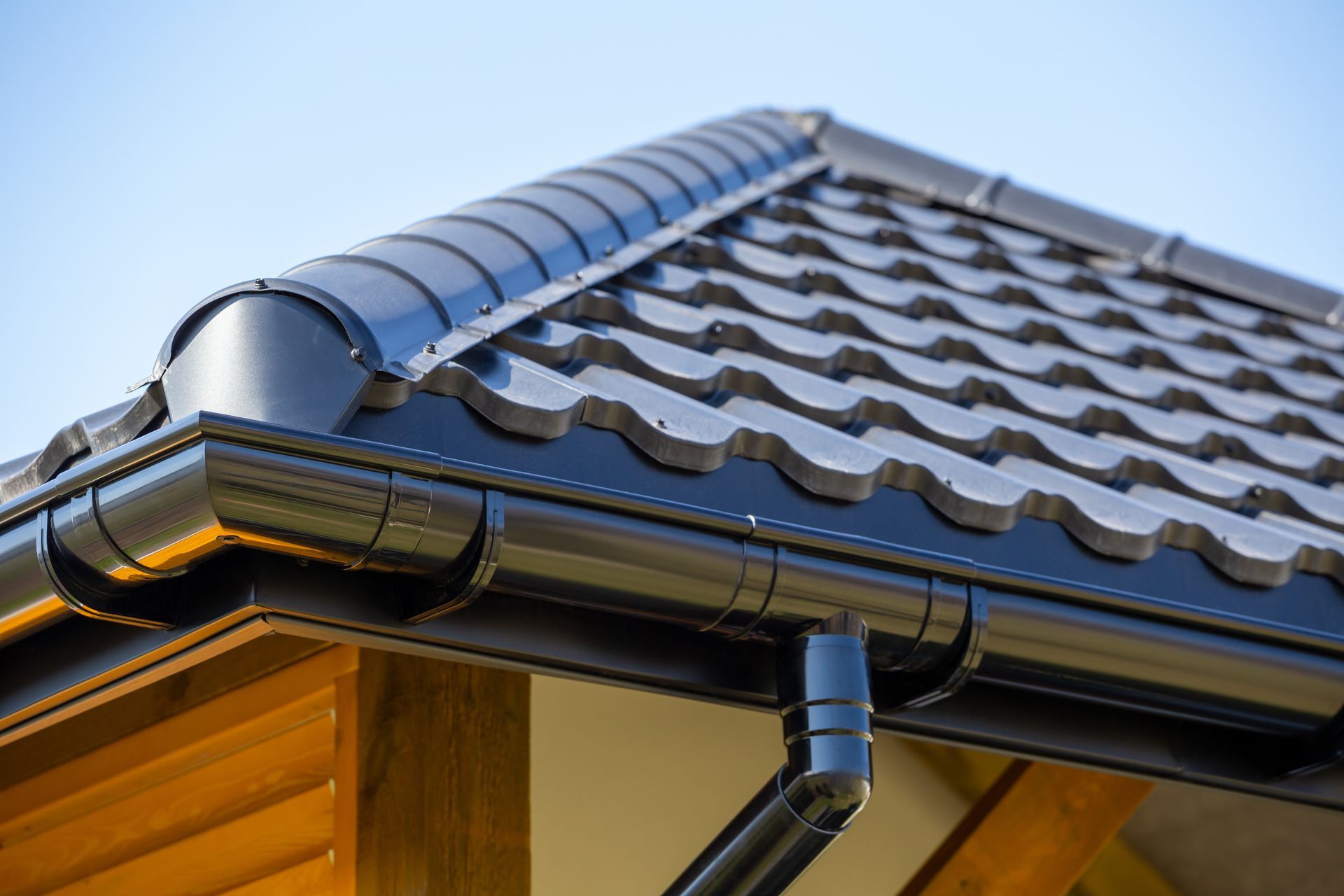 Close-up of a house roof with dark shingles and black rain gutters against a blue sky