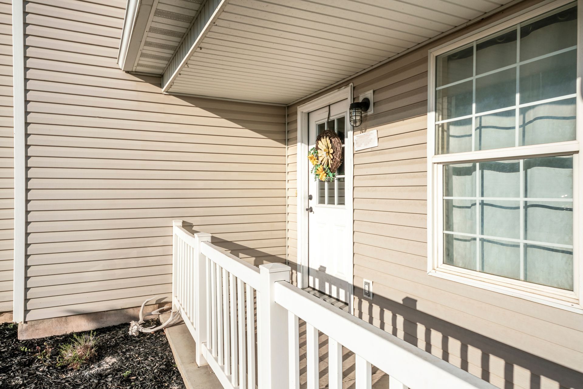 Porch of a home with tan vinyl siding, a white door with a sunflower wreath, and a white railing