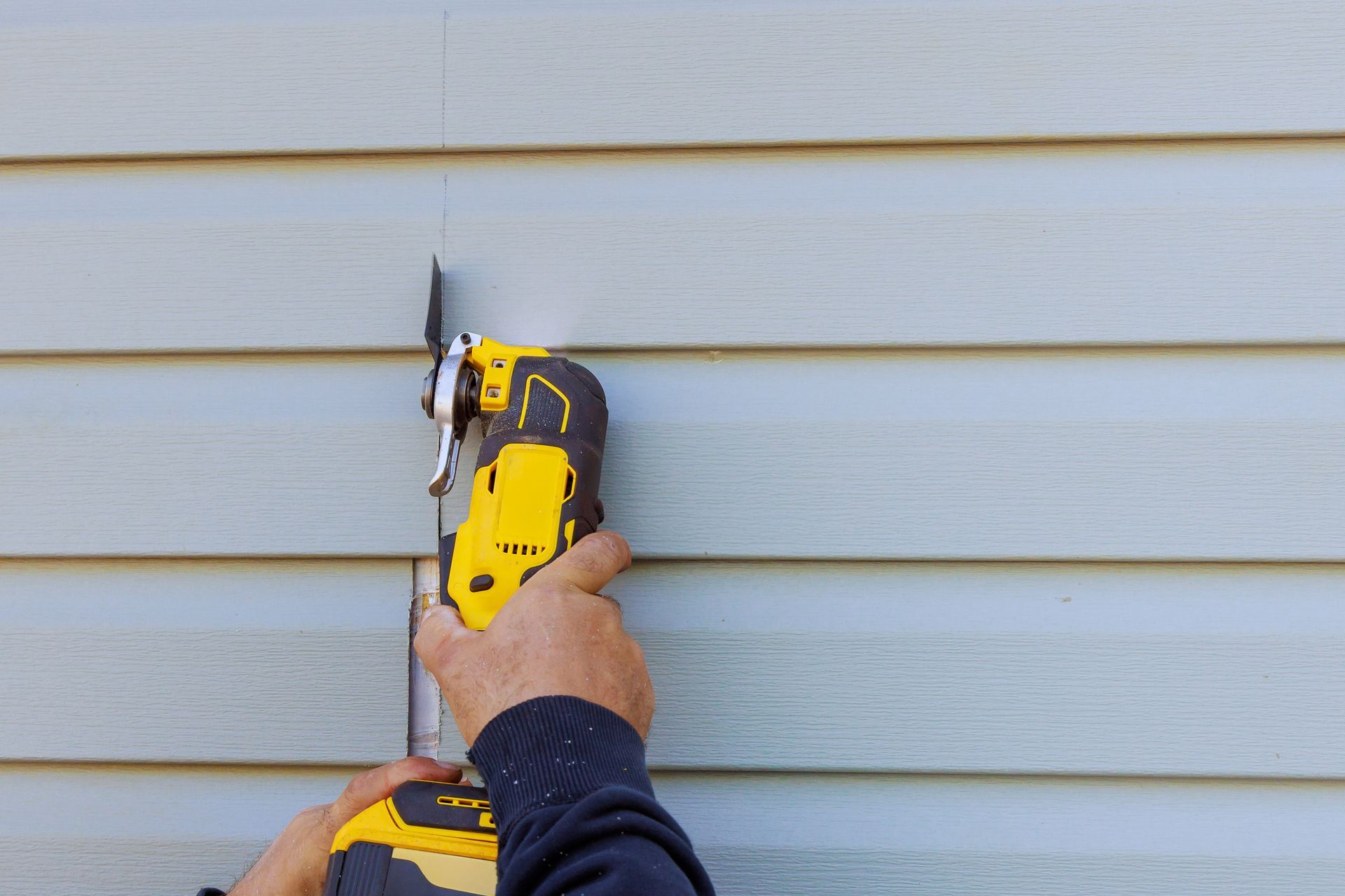 A contractor cutting a vinyl siding panel with a yellow cordless power tool