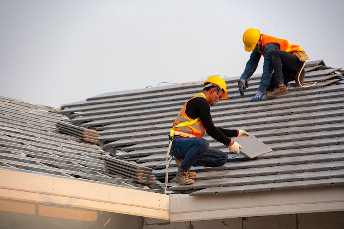 Two workers in orange safety vests and hard hats repairing a roof with gray shingles.