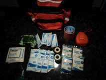 A first aid kit bag open on a dark counter, surrounded by supplies including gauze, bandages, tape, and a medical device.