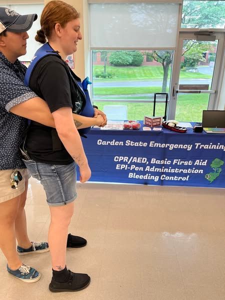 A person practices the Heimlich maneuver on another wearing a training vest in front of a first aid training table.