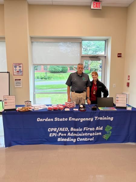 Two instructors stand behind a blue table for Garden State Emergency Training, promoting CPR and first aid classes.