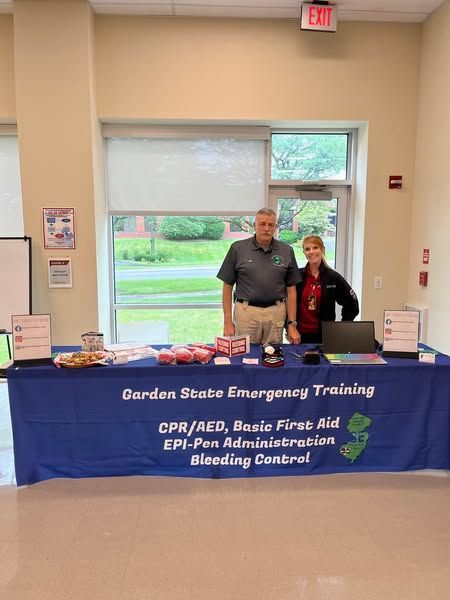 Two people stand behind a blue Garden State Emergency Training table offering first aid and CPR services in a lobby.