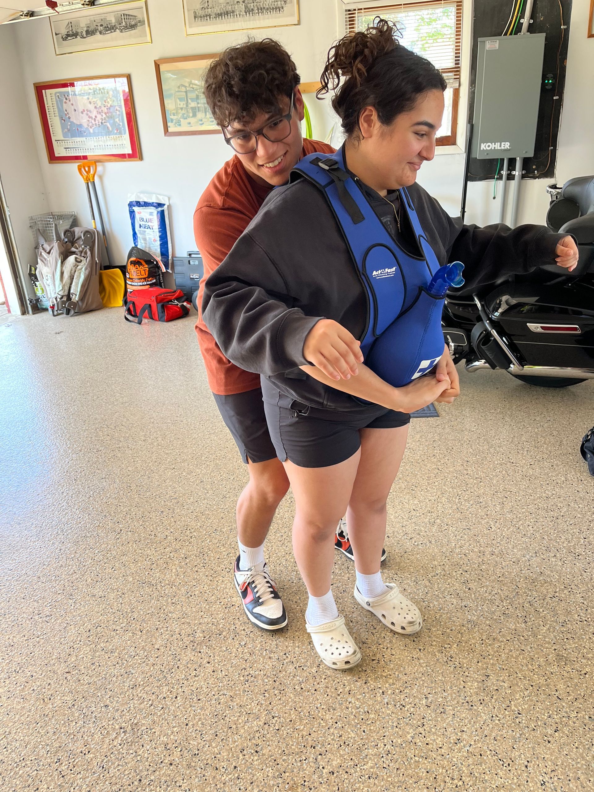 A person helping another fit a blue life vest in a garage, both smiling.