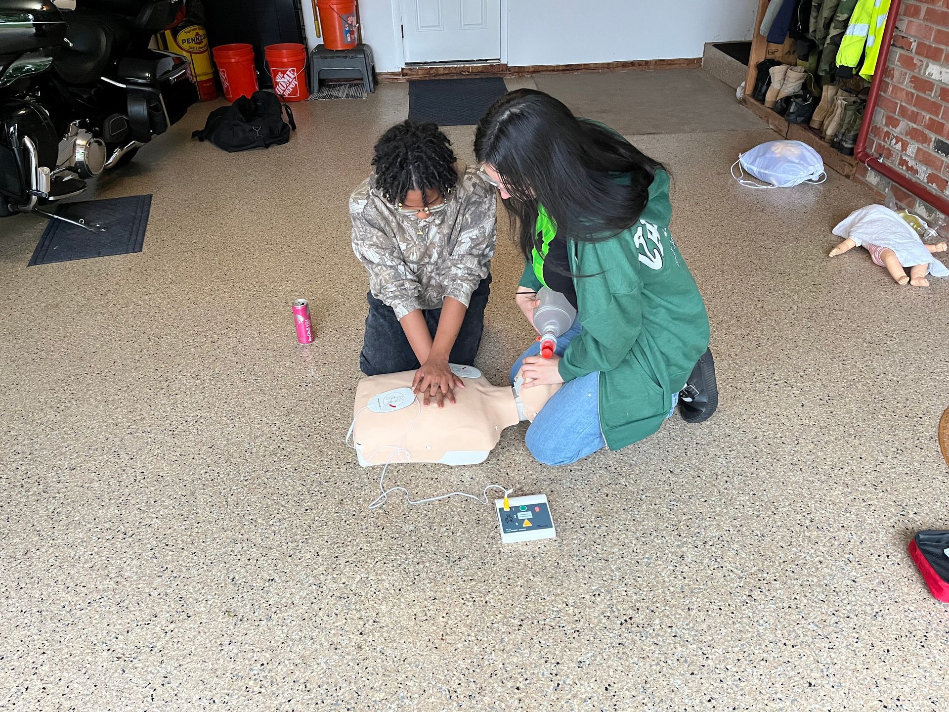 Two people practicing CPR on a training mannequin in a garage with motorcycles and equipment visible in the background.