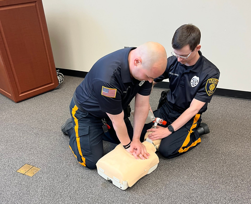 Two uniformed individuals perform CPR training on a mannequin in an indoor setting.