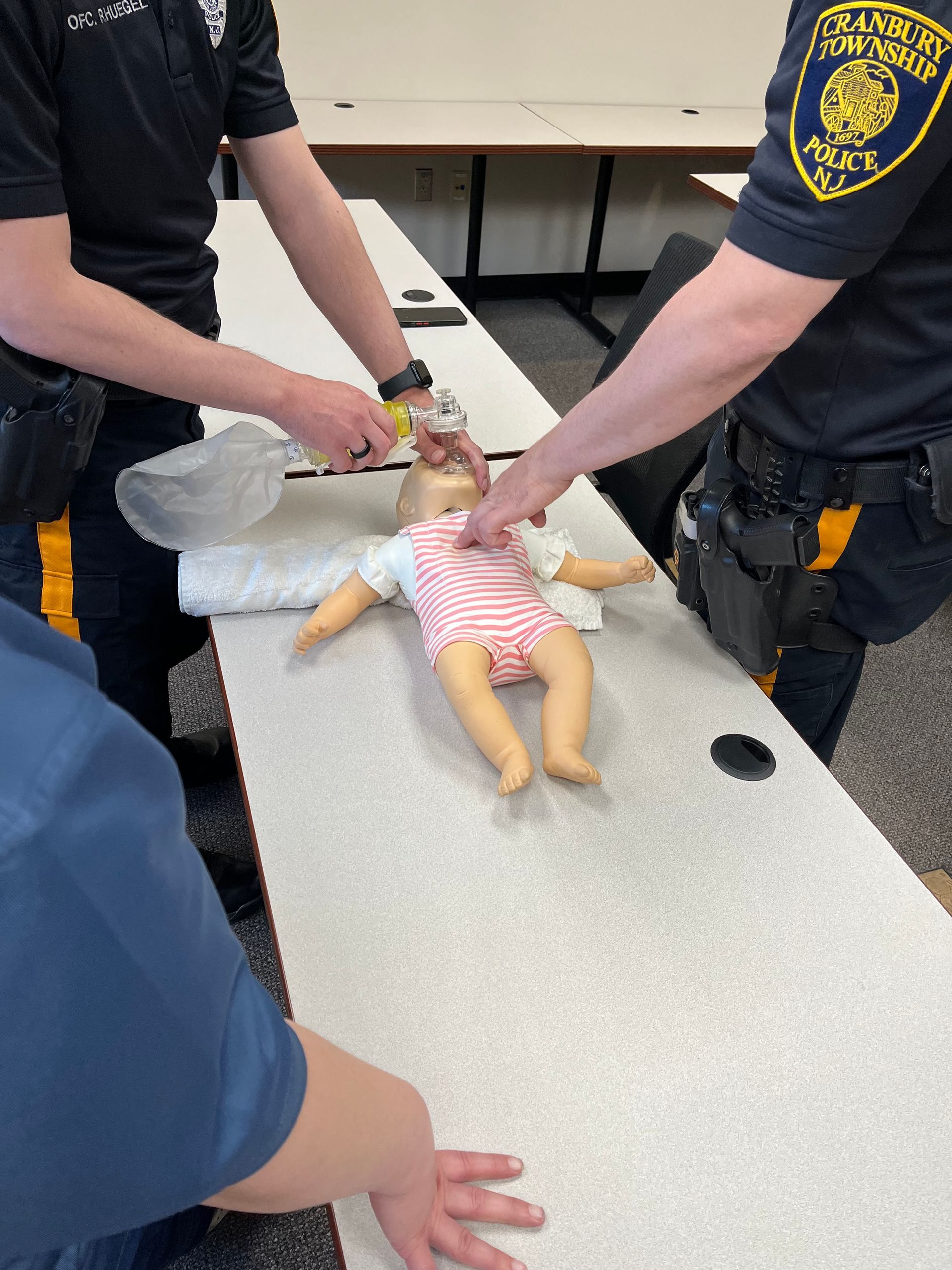 Two police officers demonstrate CPR training on an infant mannequin using a bag valve mask on a table.