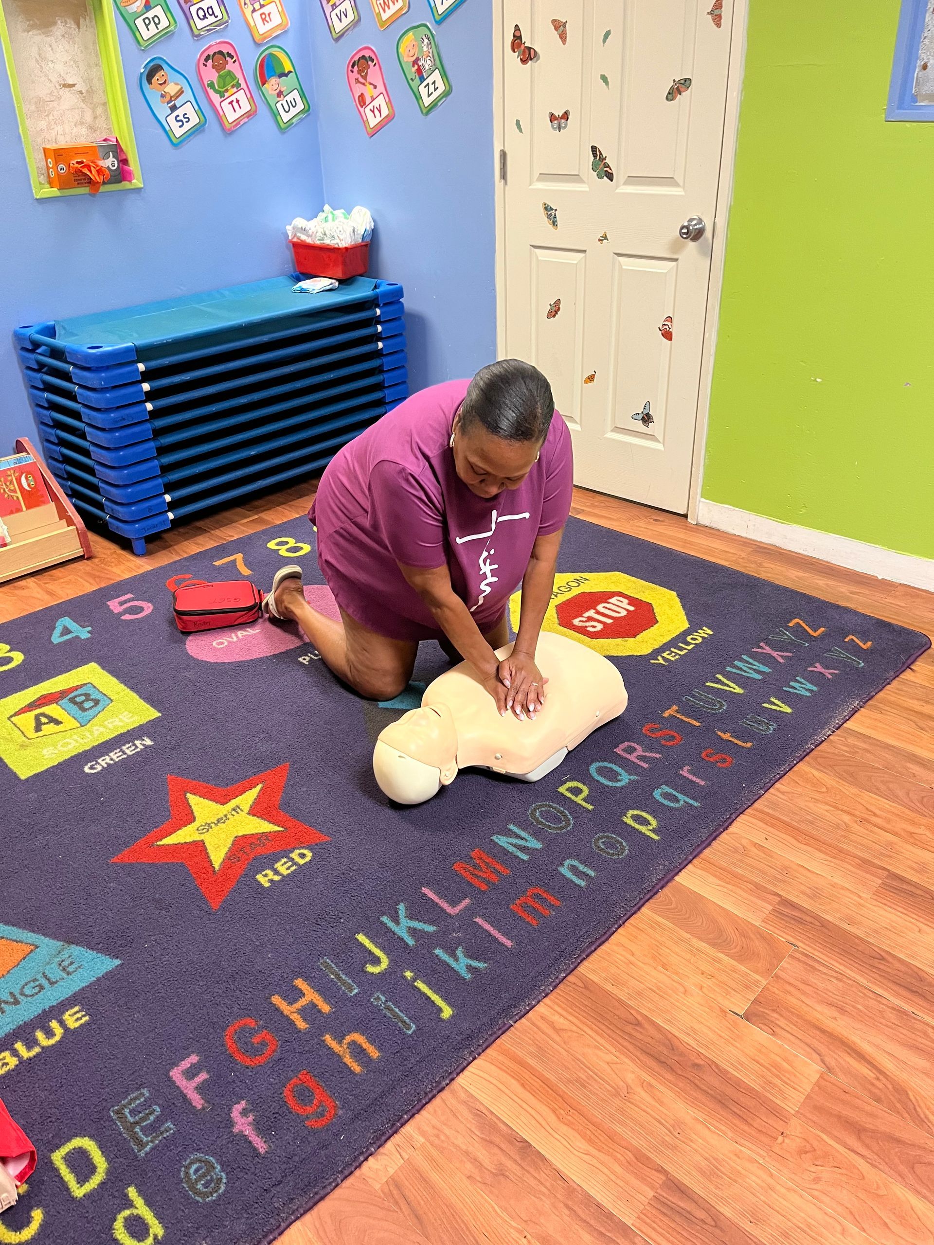 A person kneels on a colorful classroom rug, practicing CPR on an infant-sized mannequin.