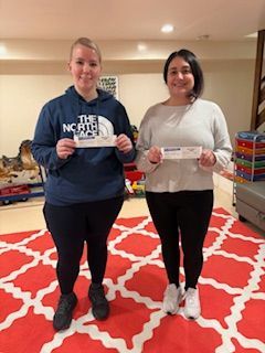 Two people stand on a patterned rug, smiling and holding small slips of paper in a room with a toy storage unit.