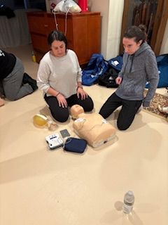 Two people kneel on a floor during a CPR training class, positioned around a mannequin and a portable defibrillator.