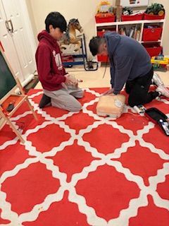 Two people practice CPR on a training mannequin on a red and white patterned rug in a playroom.