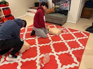 Two people kneel on a patterned red rug practicing CPR techniques on medical training mannequins.