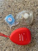A red Ambu brand CPR pocket mask case, a transparent resuscitation mask, and a blue wrapped alcohol wipe on a counter.