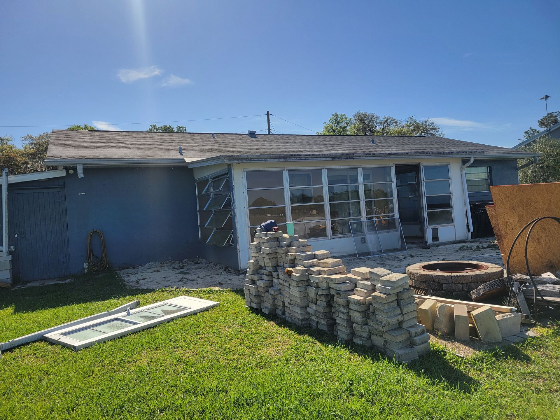 Backyard patio with stacked stone walls, sliding glass doors, and a ladder on the lawn under a blue sky