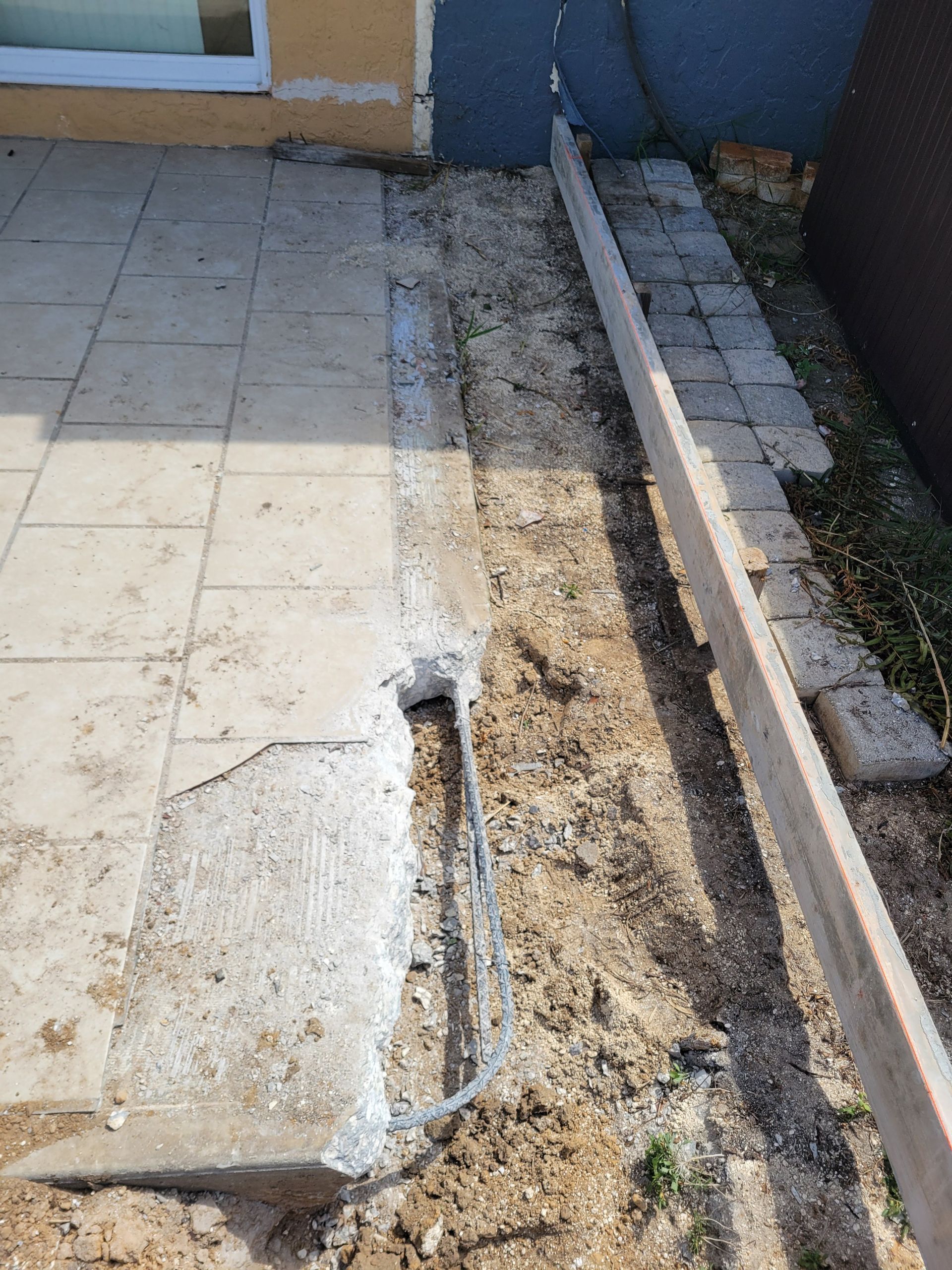 Broken concrete walkway beside a building, with a long trench and uneven pavers.