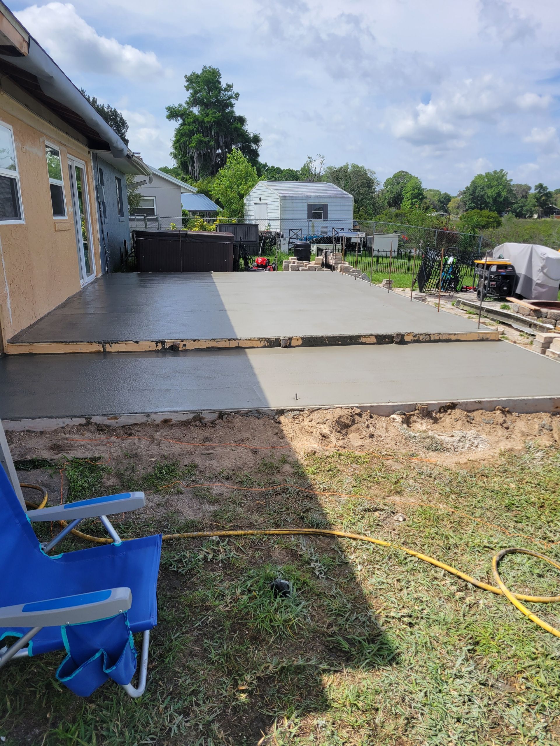 Backyard concrete patio beside a house, with chairs, tools, and a yellow hose on the grass.