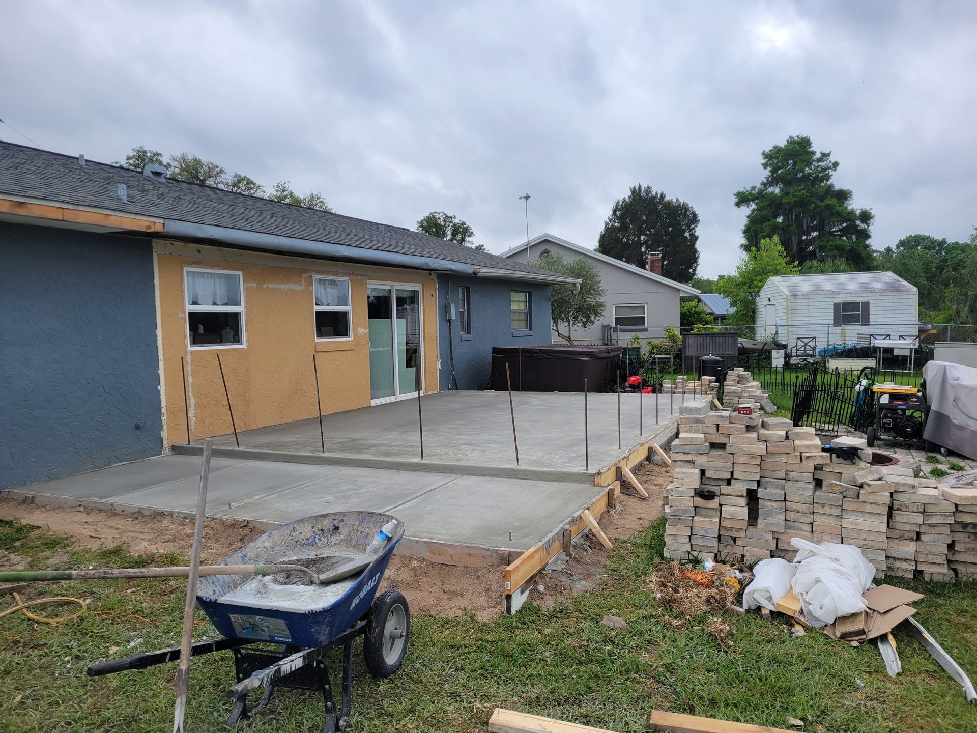 Backyard concrete patio under construction beside a blue house, with lumber, wheelbarrow, and stacked pavers