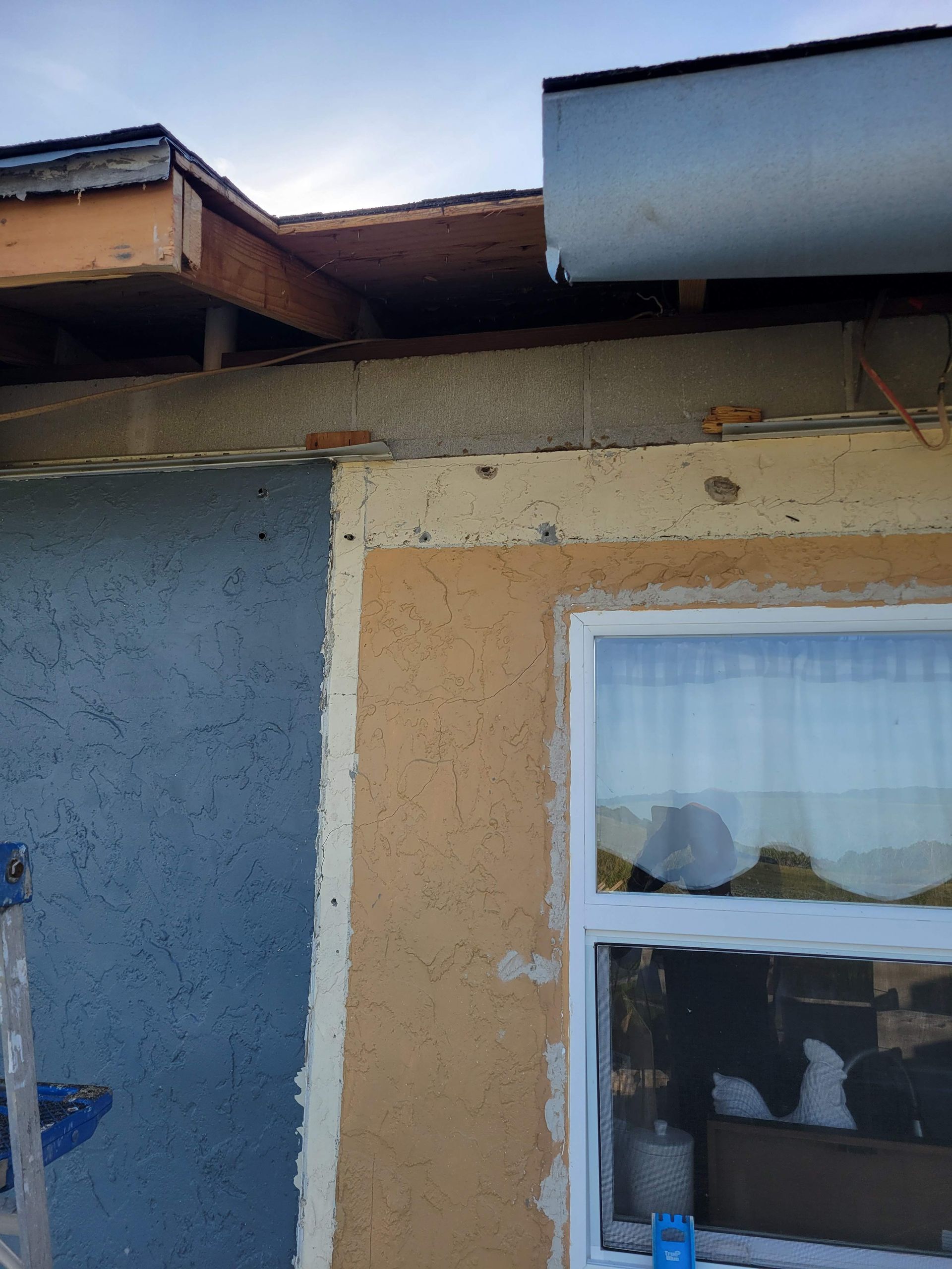 Side of a house with blue and tan stucco, a white-framed window, and roof eaves visible above.