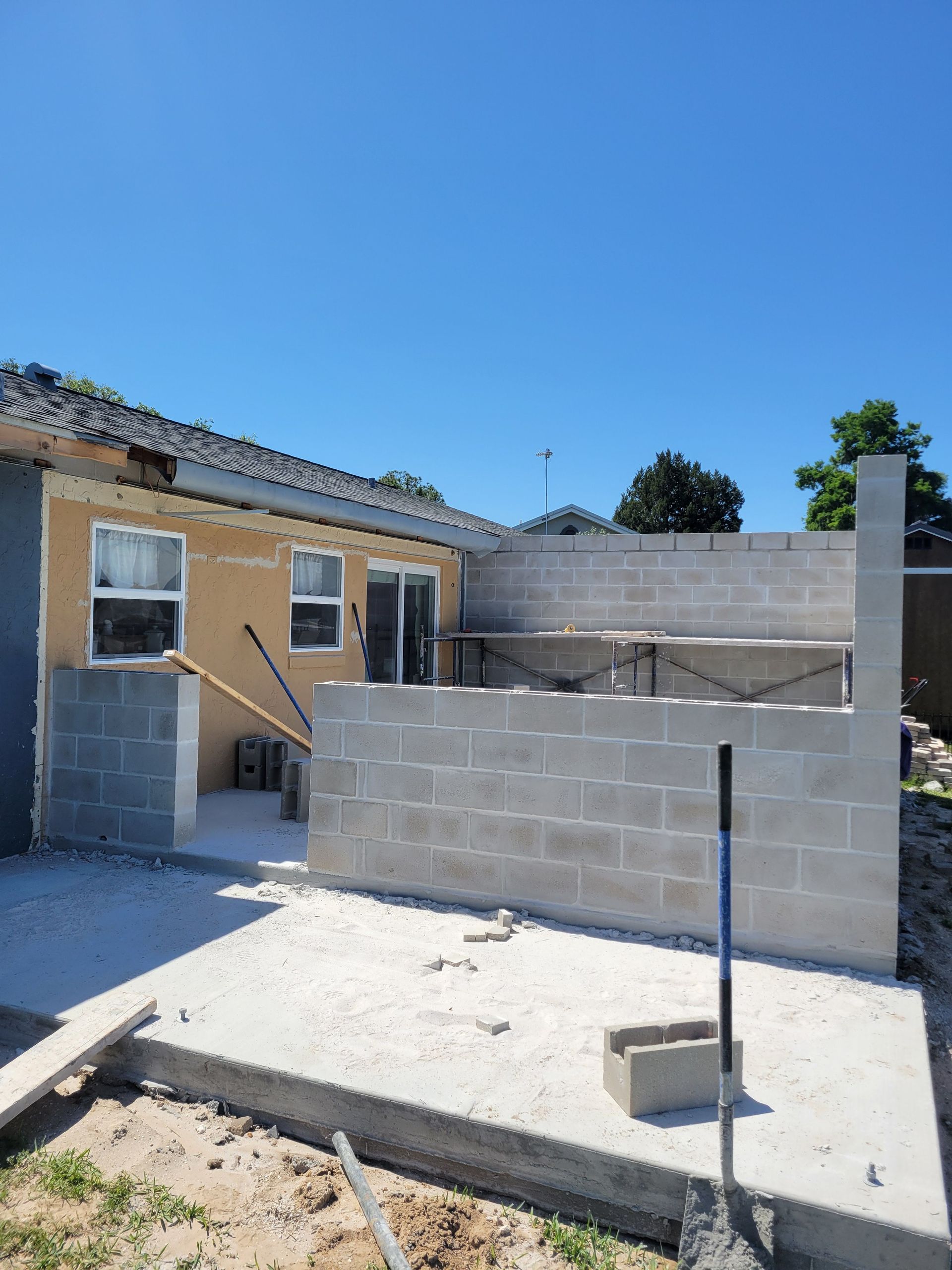 Partially built cinder block wall beside a tan house under a clear blue sky