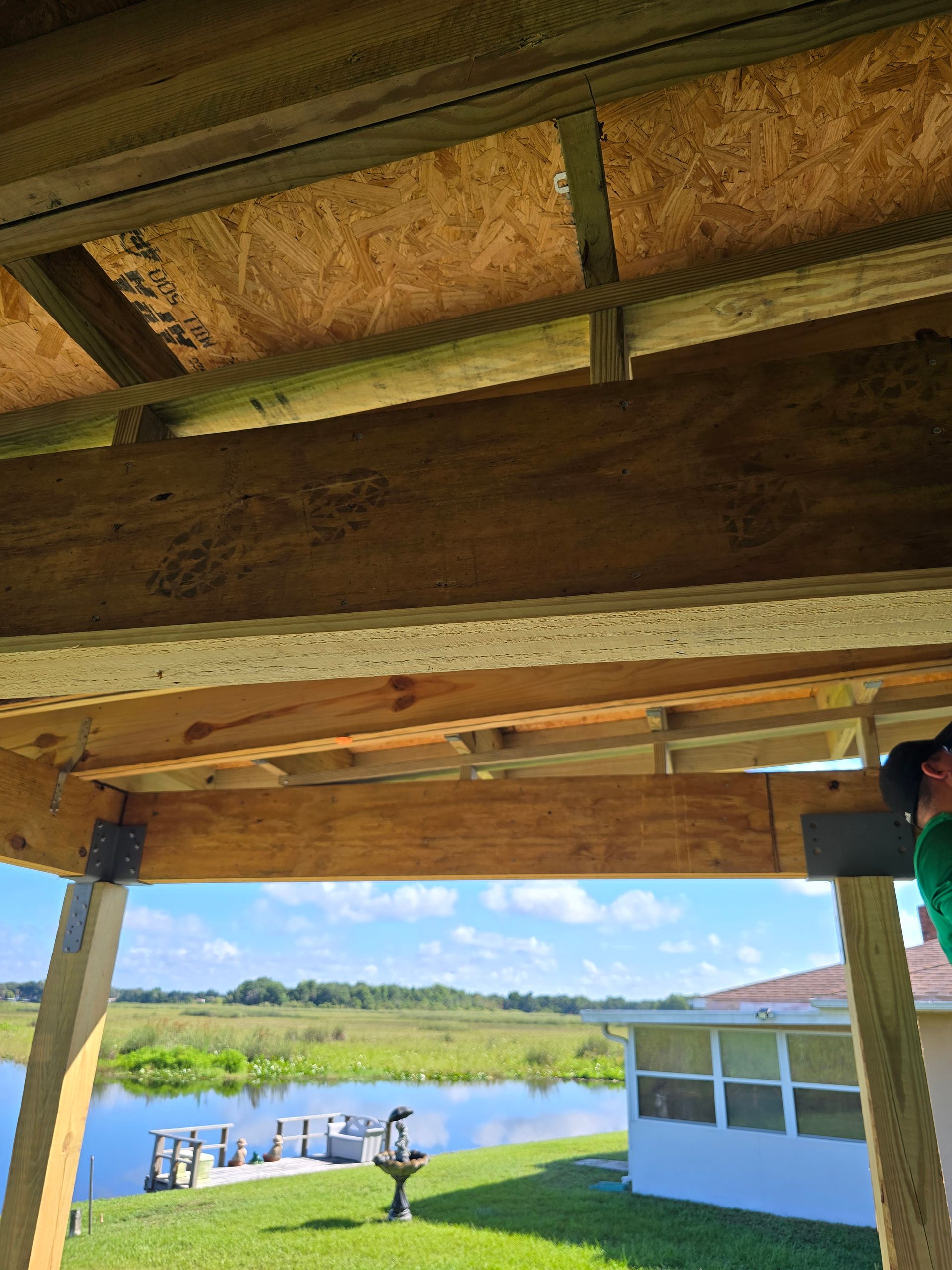 Lakefront dock view from under a wooden roof, with calm water, a small boat lift, and a white house in the distance