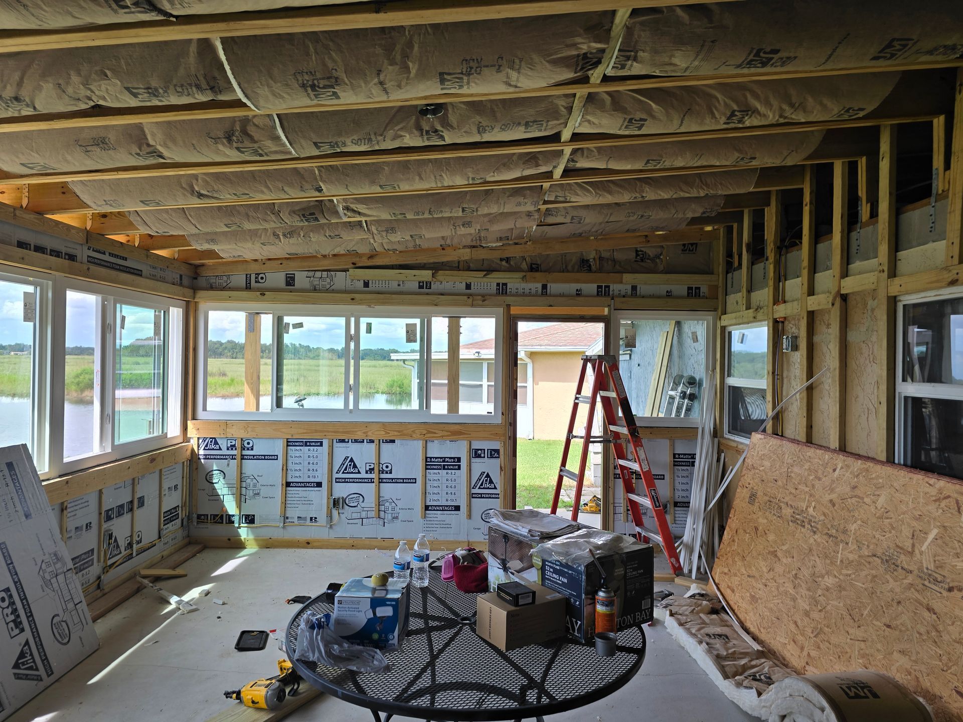 Unfinished room under construction with exposed insulation, windows, tools, and a red ladder.