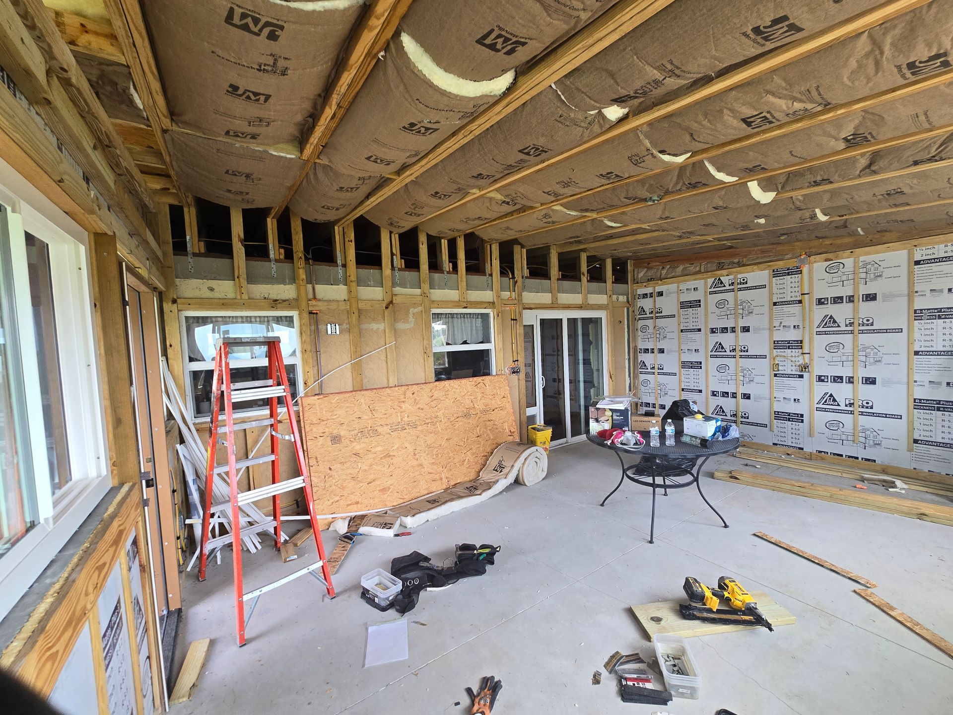 Unfinished room under renovation with exposed framing, insulation, ladders, tools, and construction materials.