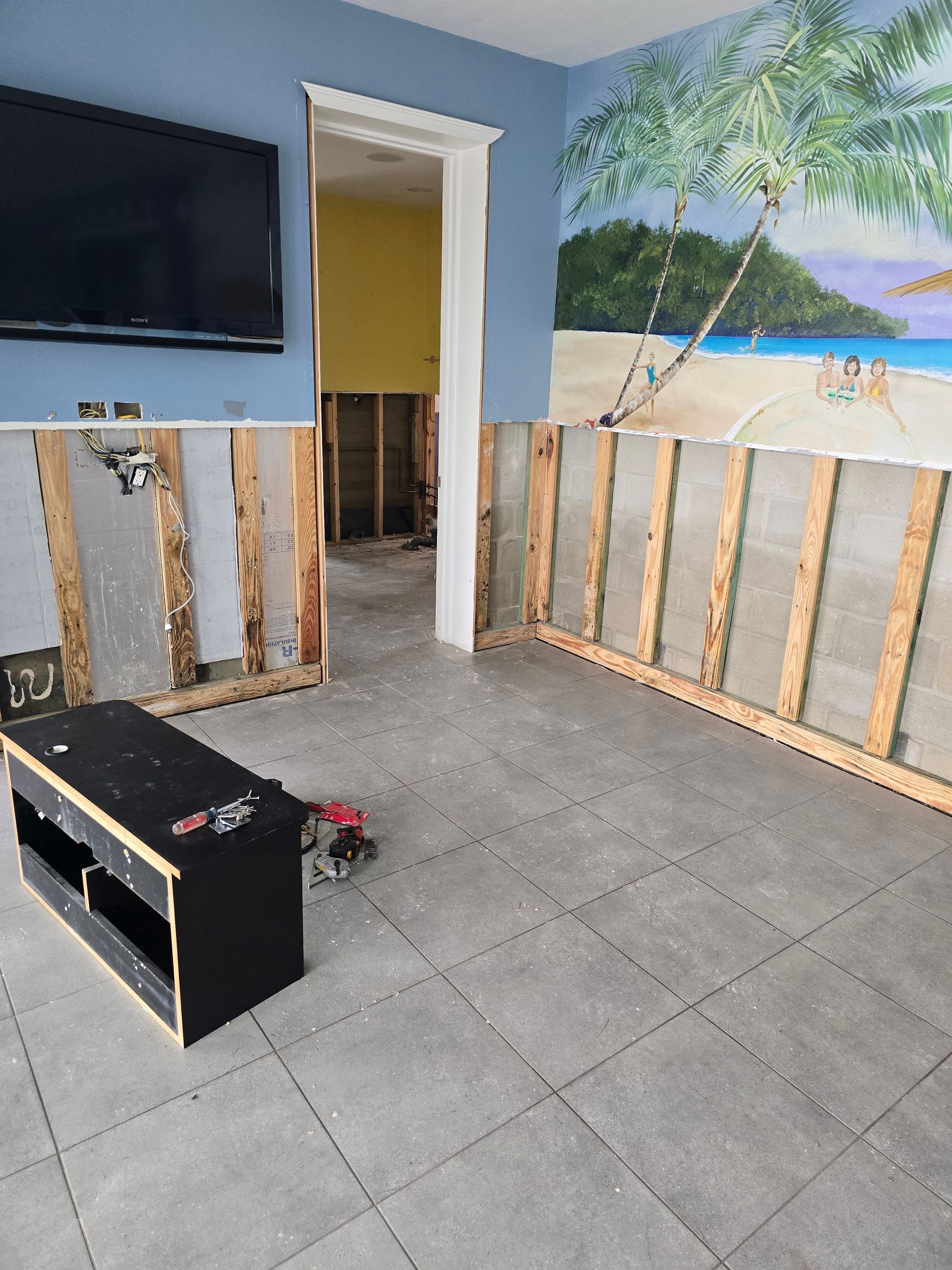 Living room under renovation with exposed wall framing, tiled floor, TV, and beach mural.
