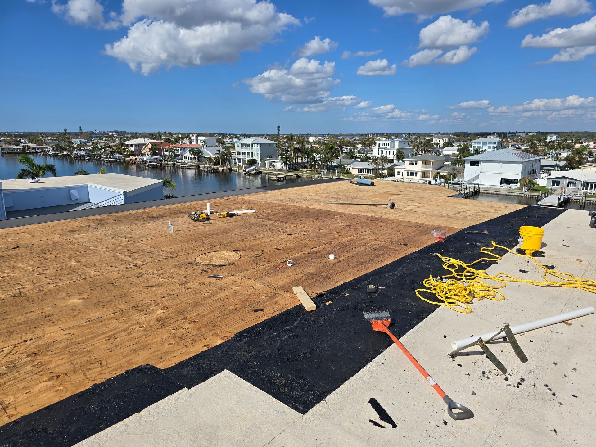 Flat rooftop under construction with exposed plywood, black underlayment, tools, and a cityscape under blue sky.