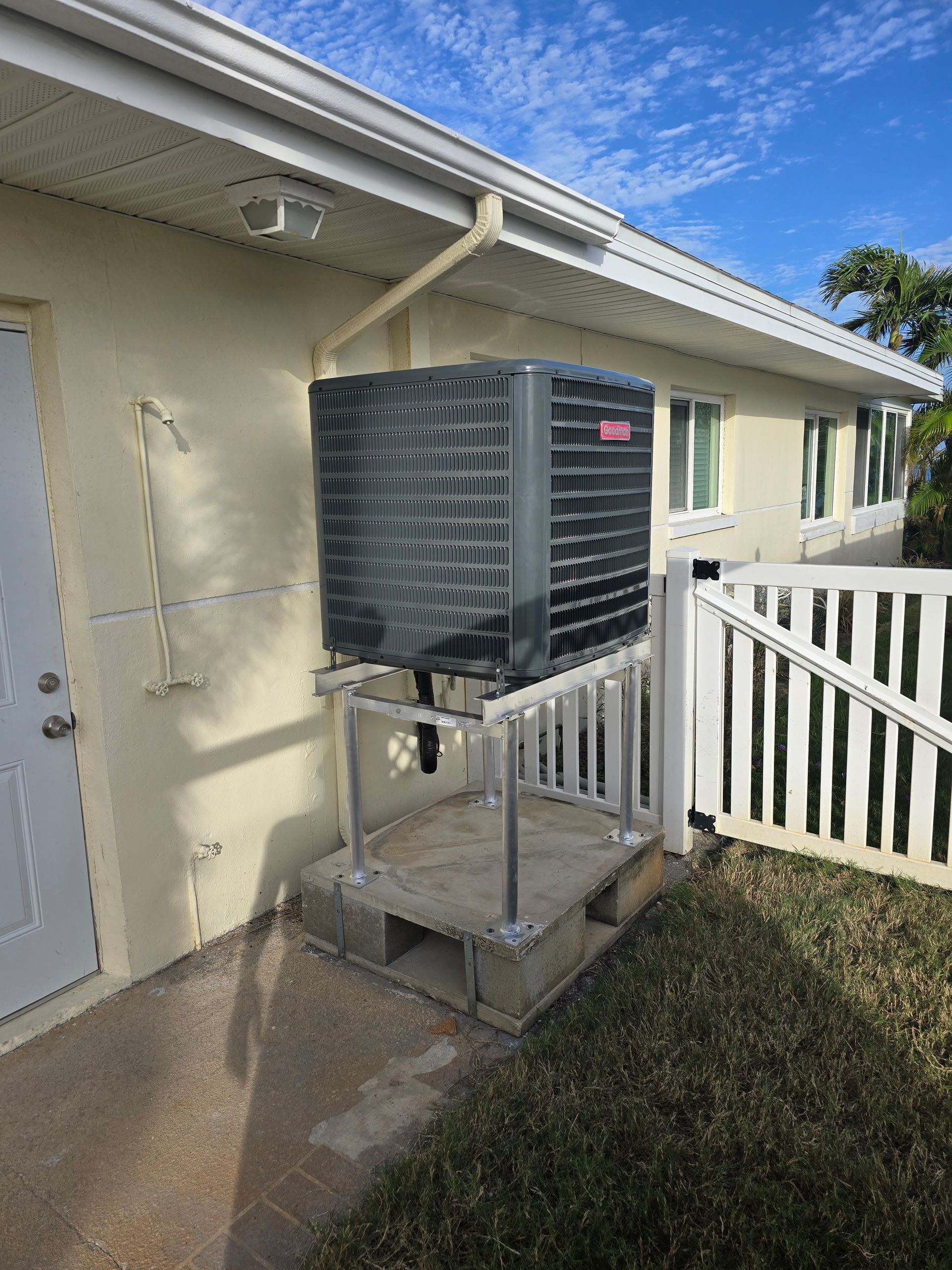 Outdoor air conditioner unit beside a beige house, mounted on a metal stand near a white railing.