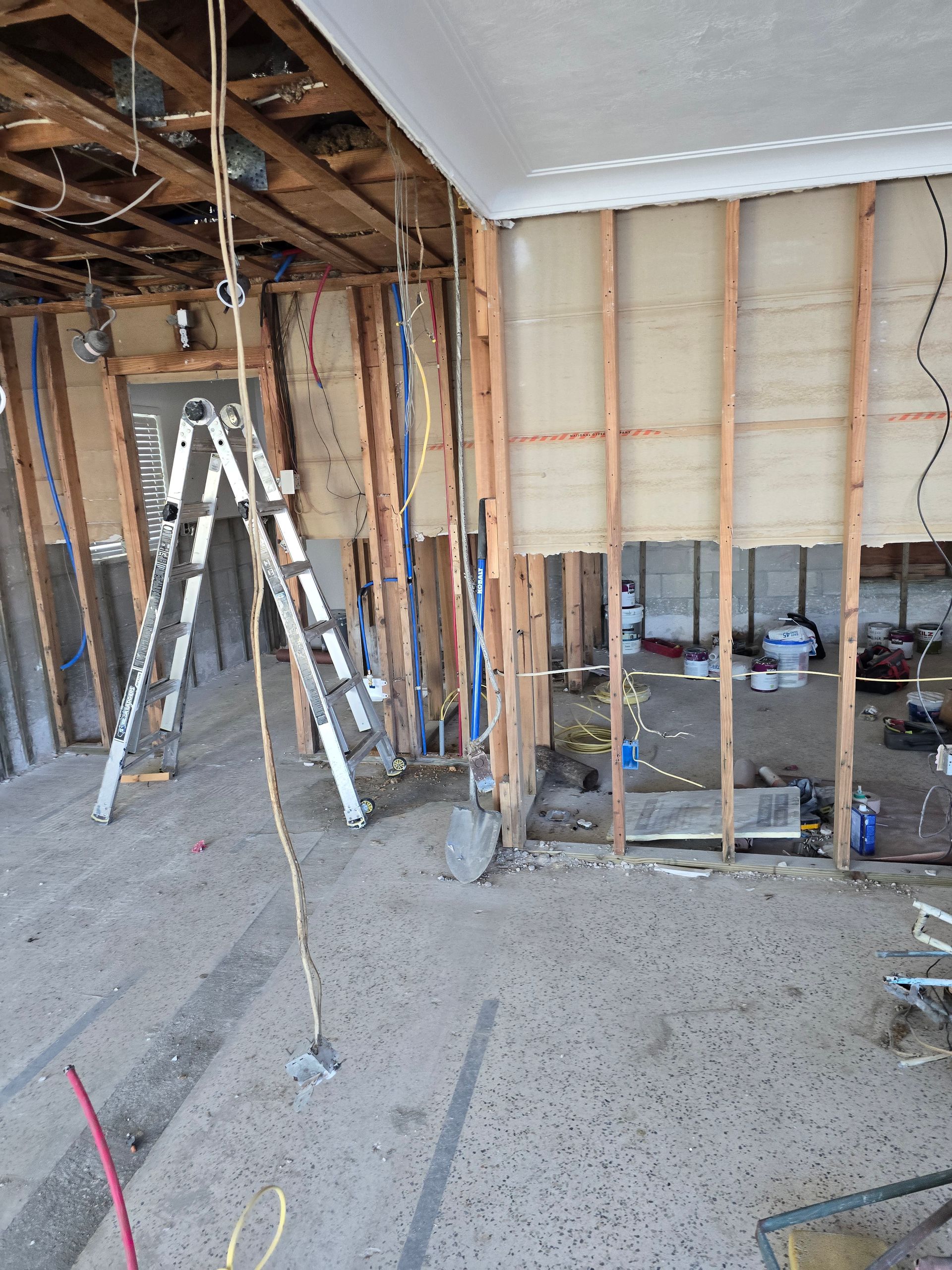 Interior under renovation with exposed studs, ladders, wiring, and unfinished flooring