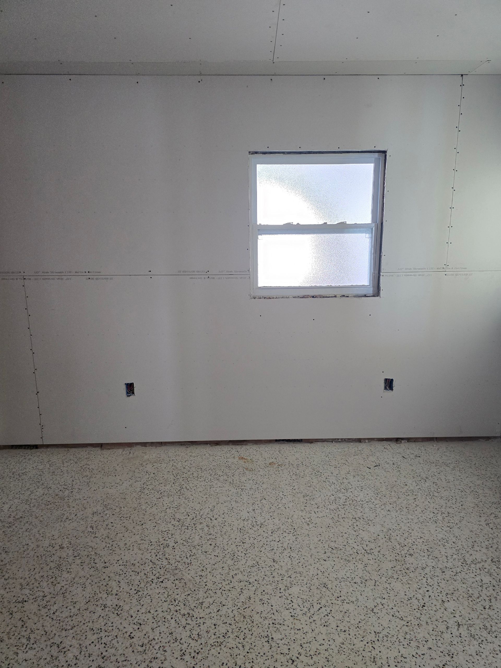 Empty room with white walls, speckled floor, and a small window letting in bright light.