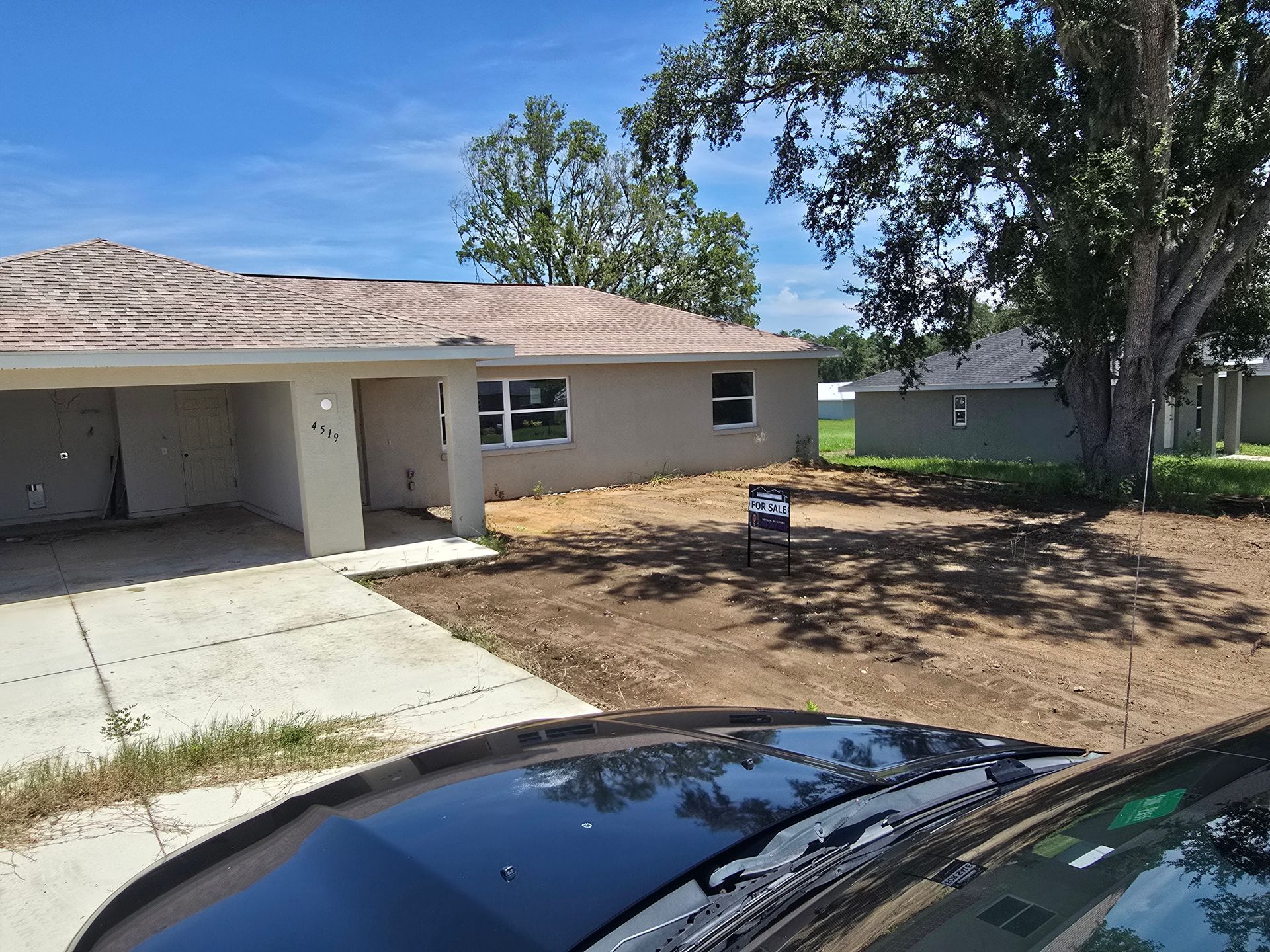 Single-story house with a driveway, bare front yard, and a car hood in the foreground.
