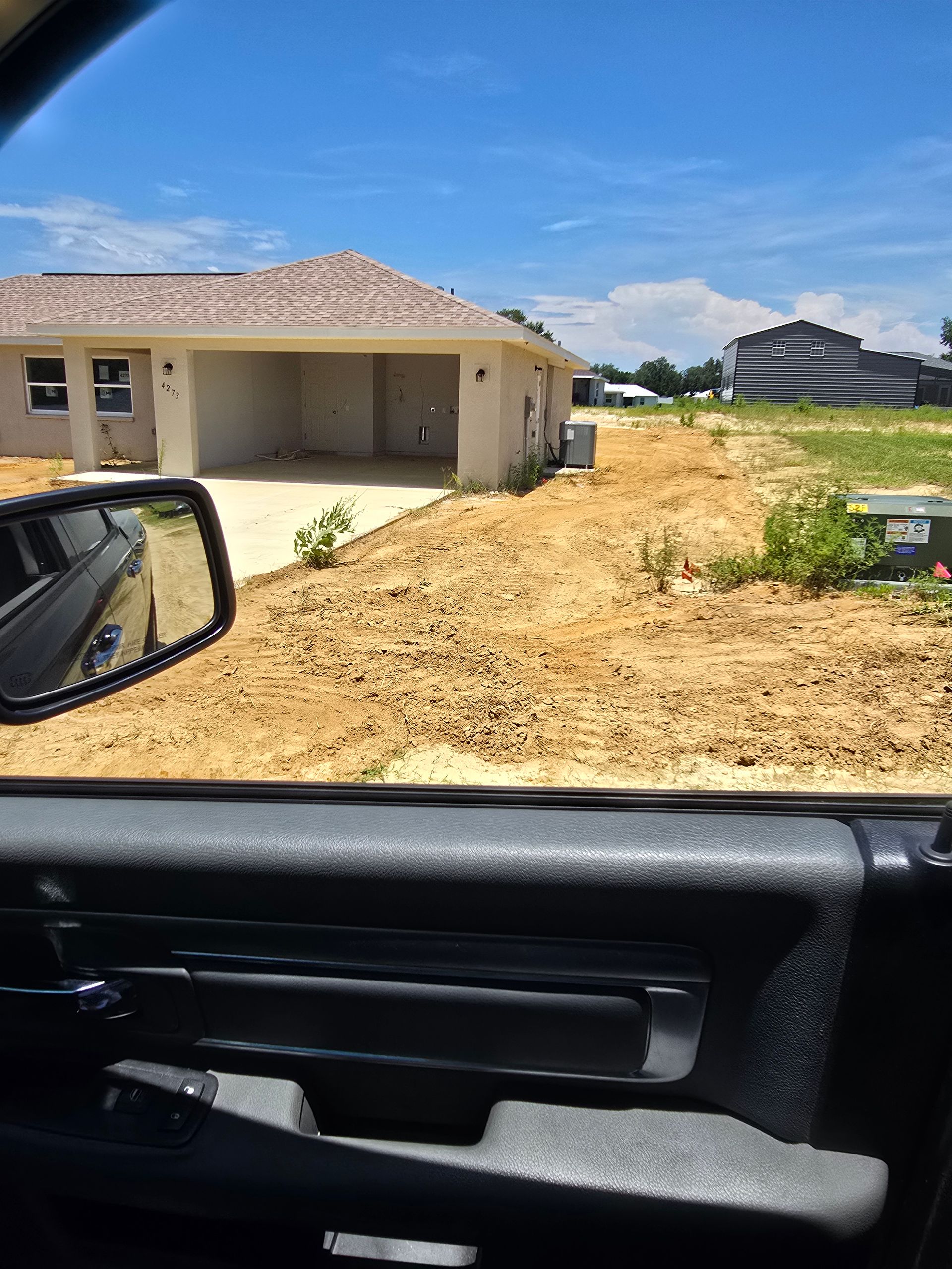View from a car of a beige house under construction beside a dirt lot on a sunny day