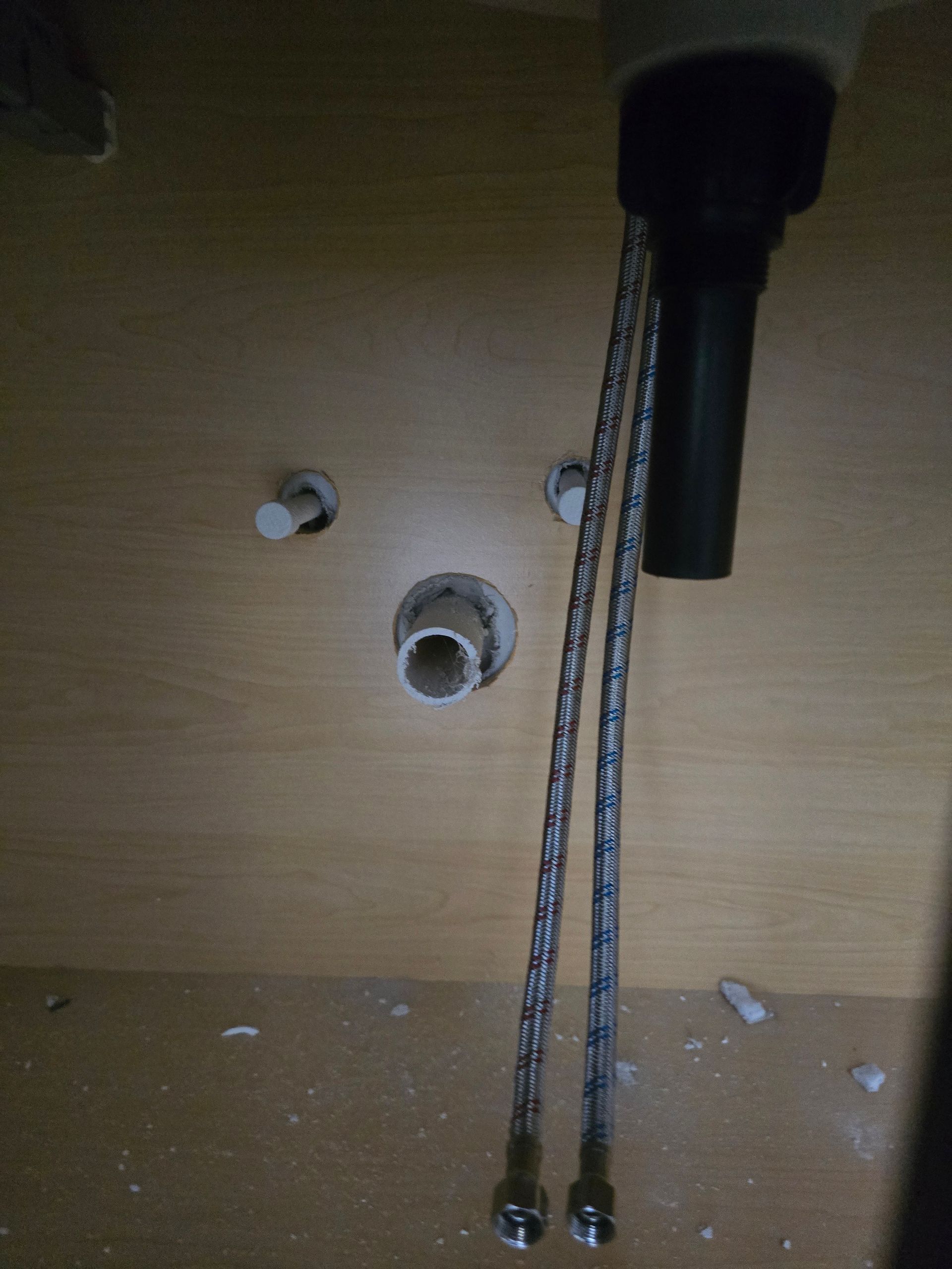 Under-sink cabinet with plumbing pipes, hoses, and wall openings against a wood panel background