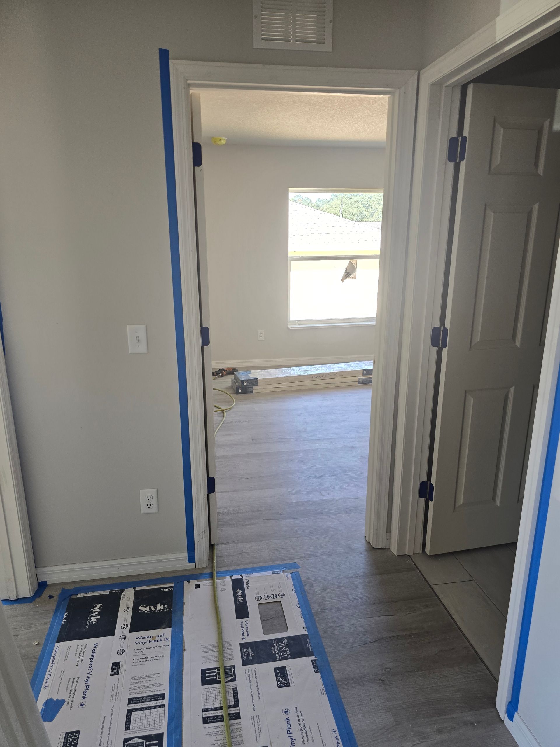 Interior hallway with blue painter’s tape around a doorway, leading to a bright room with a window.