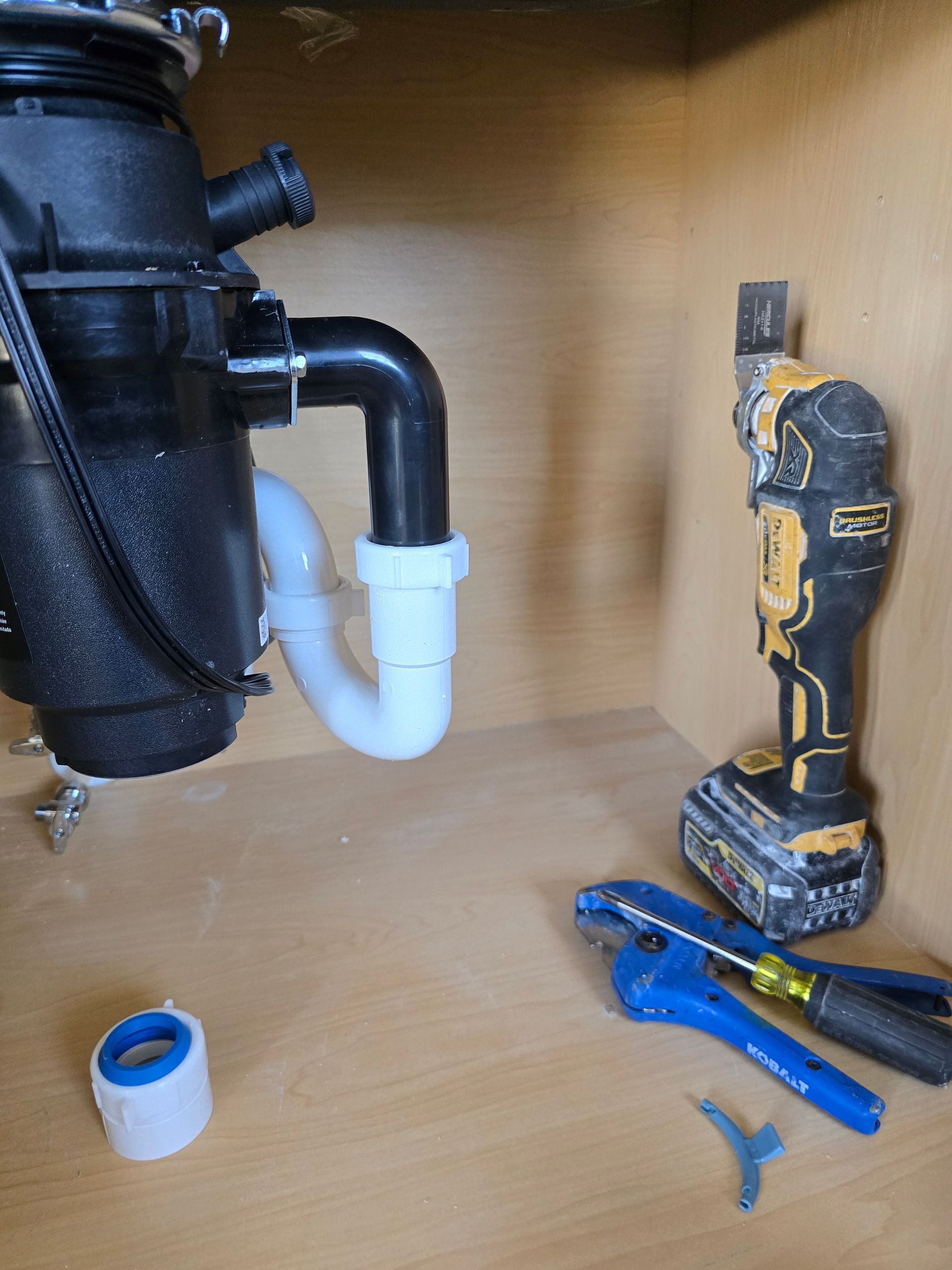 Under-sink plumbing with white PVC trap, blue wrench, cordless drill, and Teflon tape on a cabinet floor