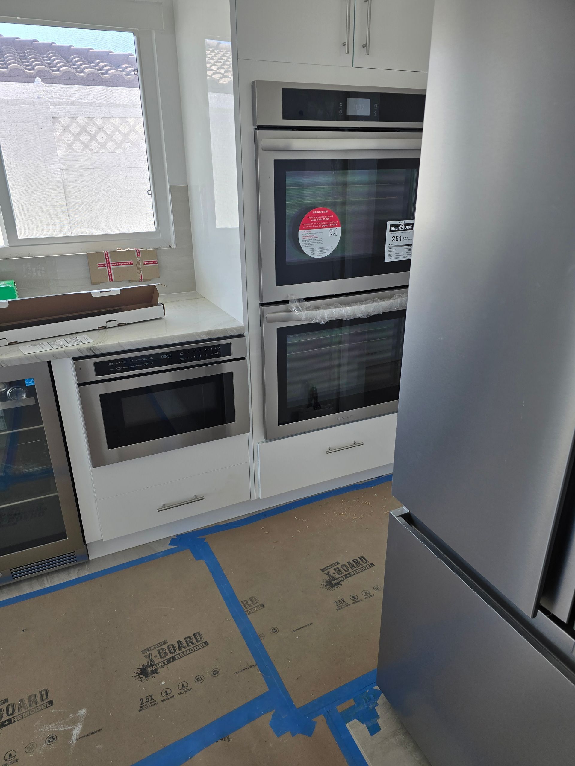 Modern kitchen with white cabinets, built-in oven, window, and appliances covered for renovation