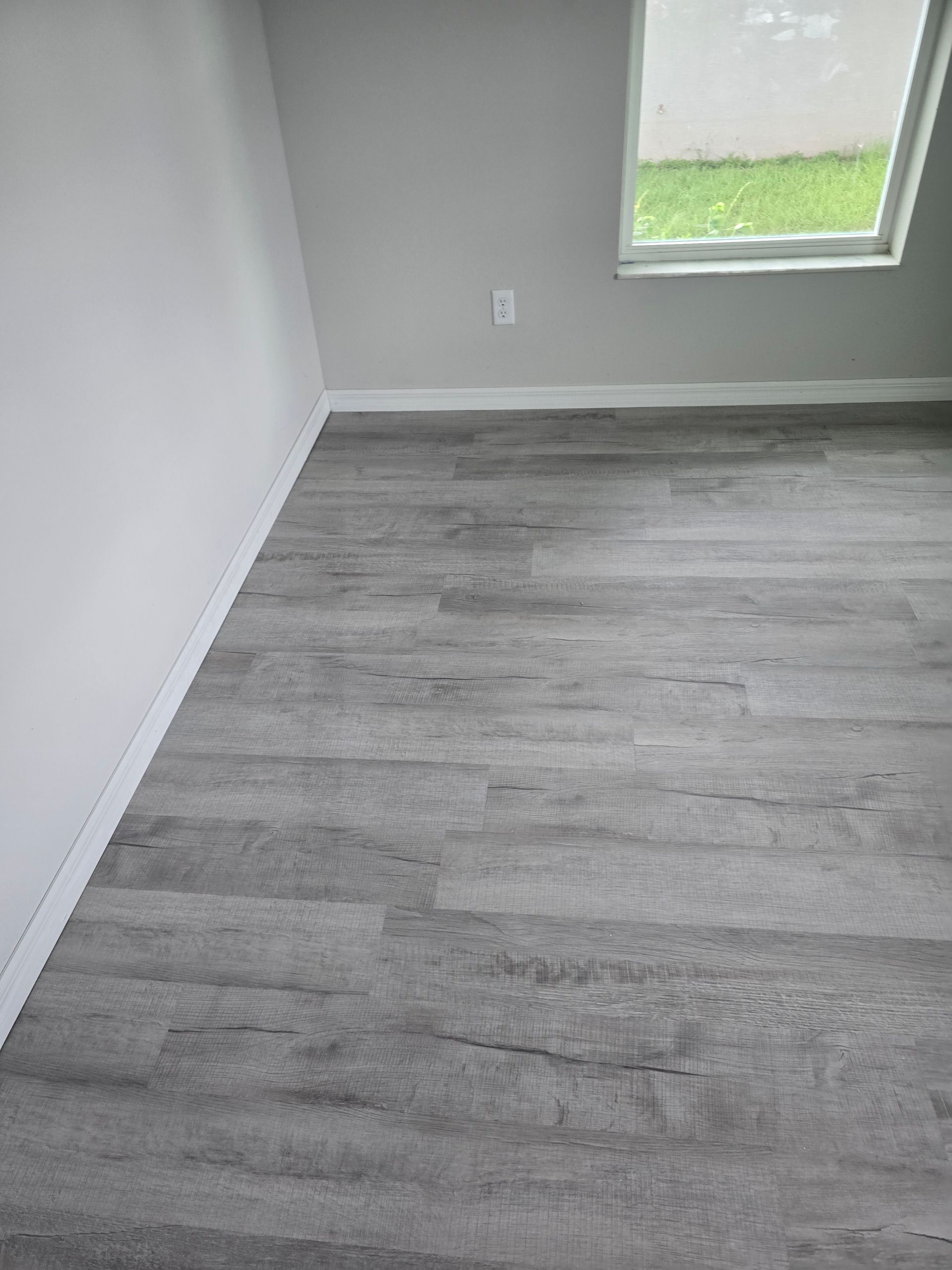 Empty room with gray wood-look flooring, white walls, and a window showing a grassy yard.