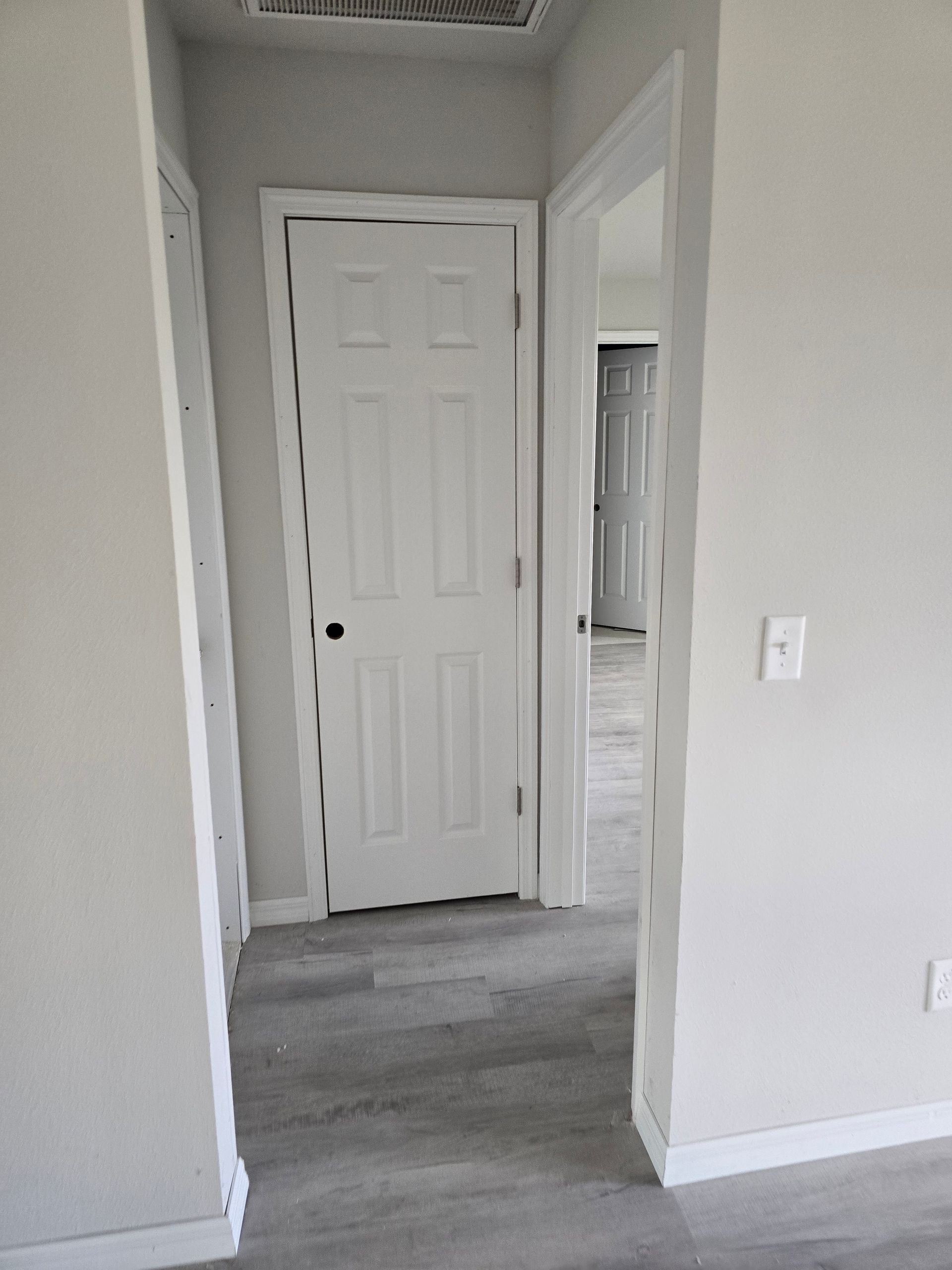 White hallway with a closed closet door, gray carpet, and a doorway leading to a brighter room.