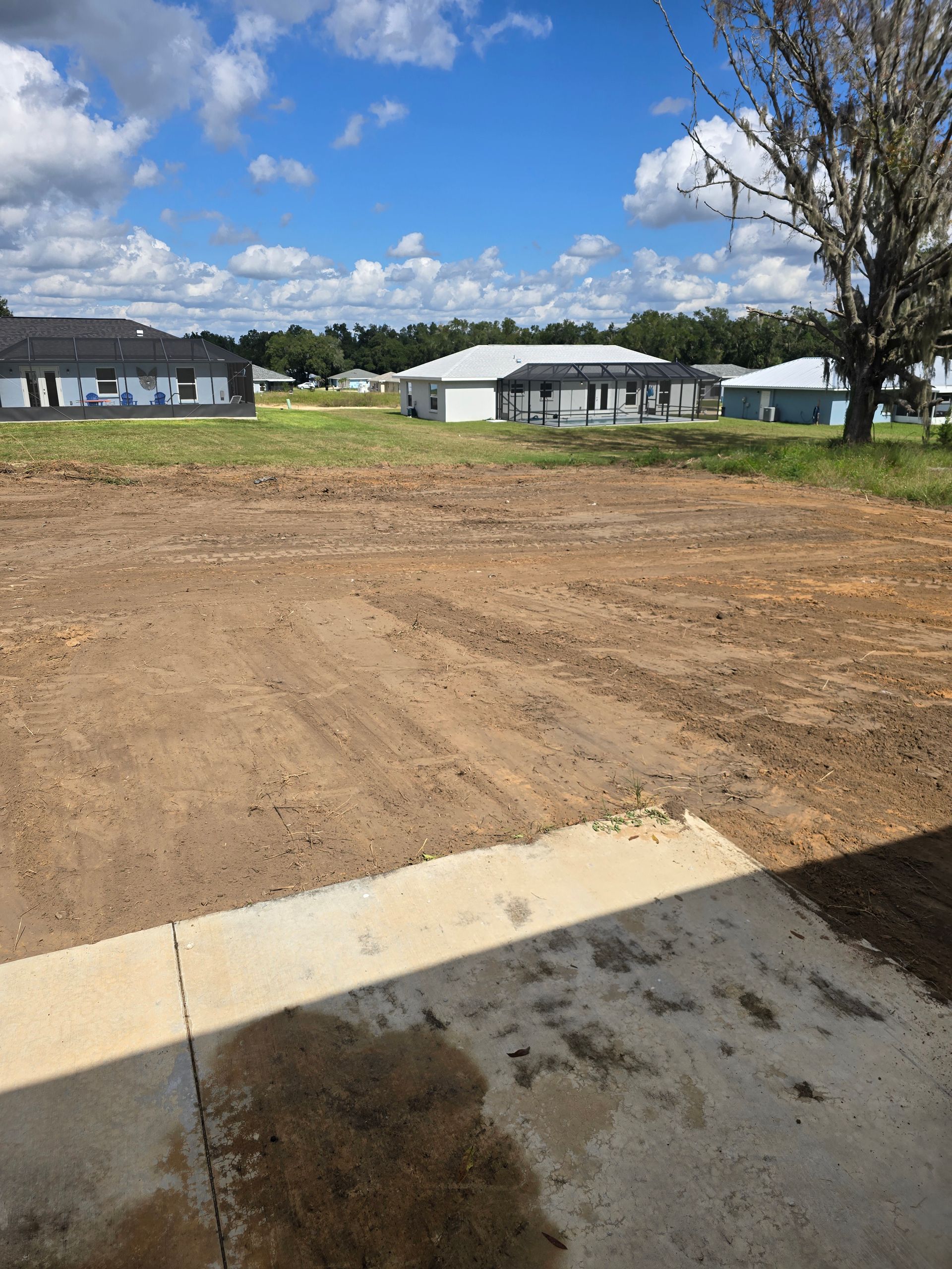 Dirt lot under blue sky with scattered clouds, concrete edge in foreground, and houses in the distance.